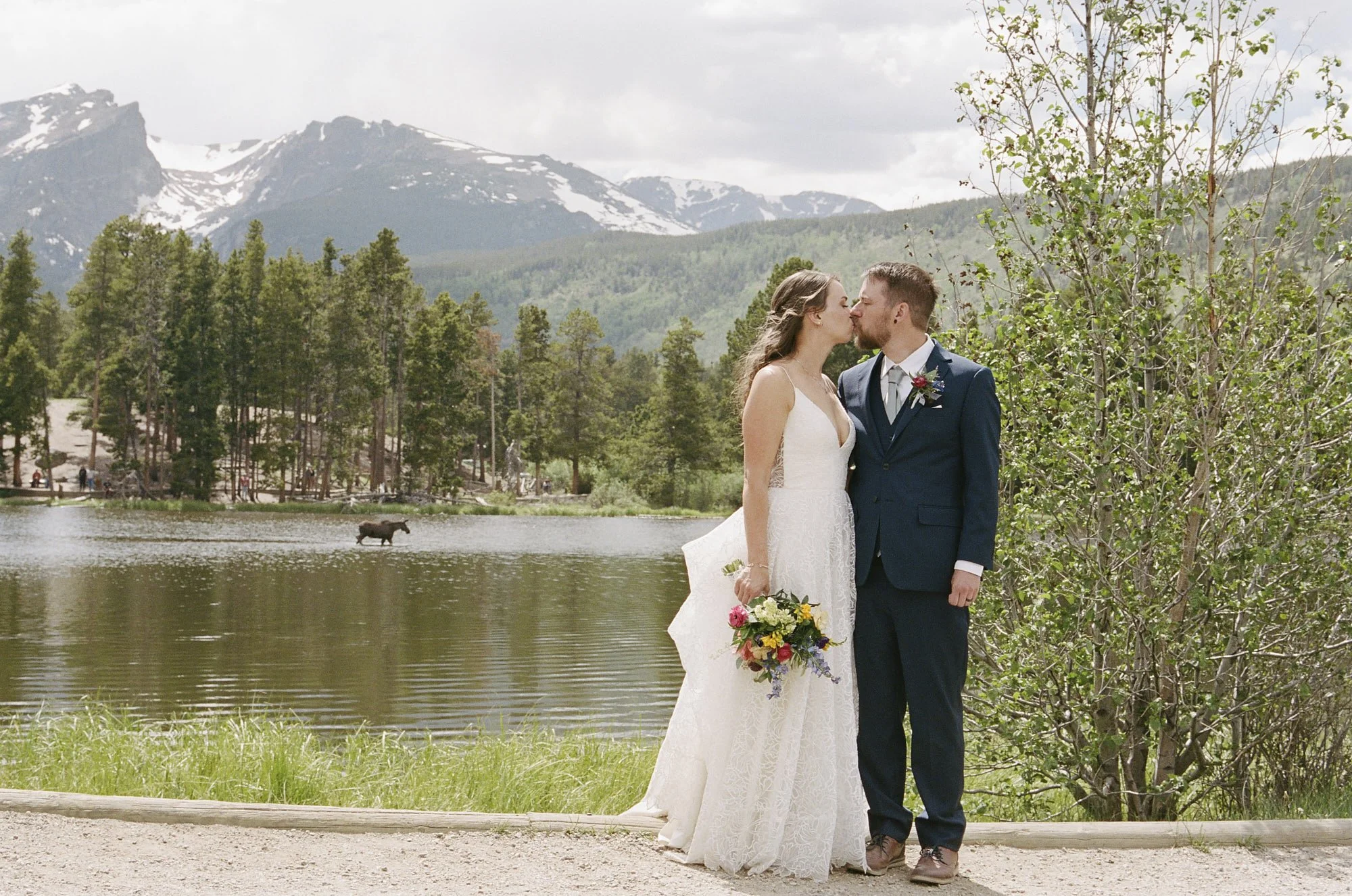 A 35mm film photo of a couple kissing in front of a lake in Rocky Mountain National Park with a moose behind them.