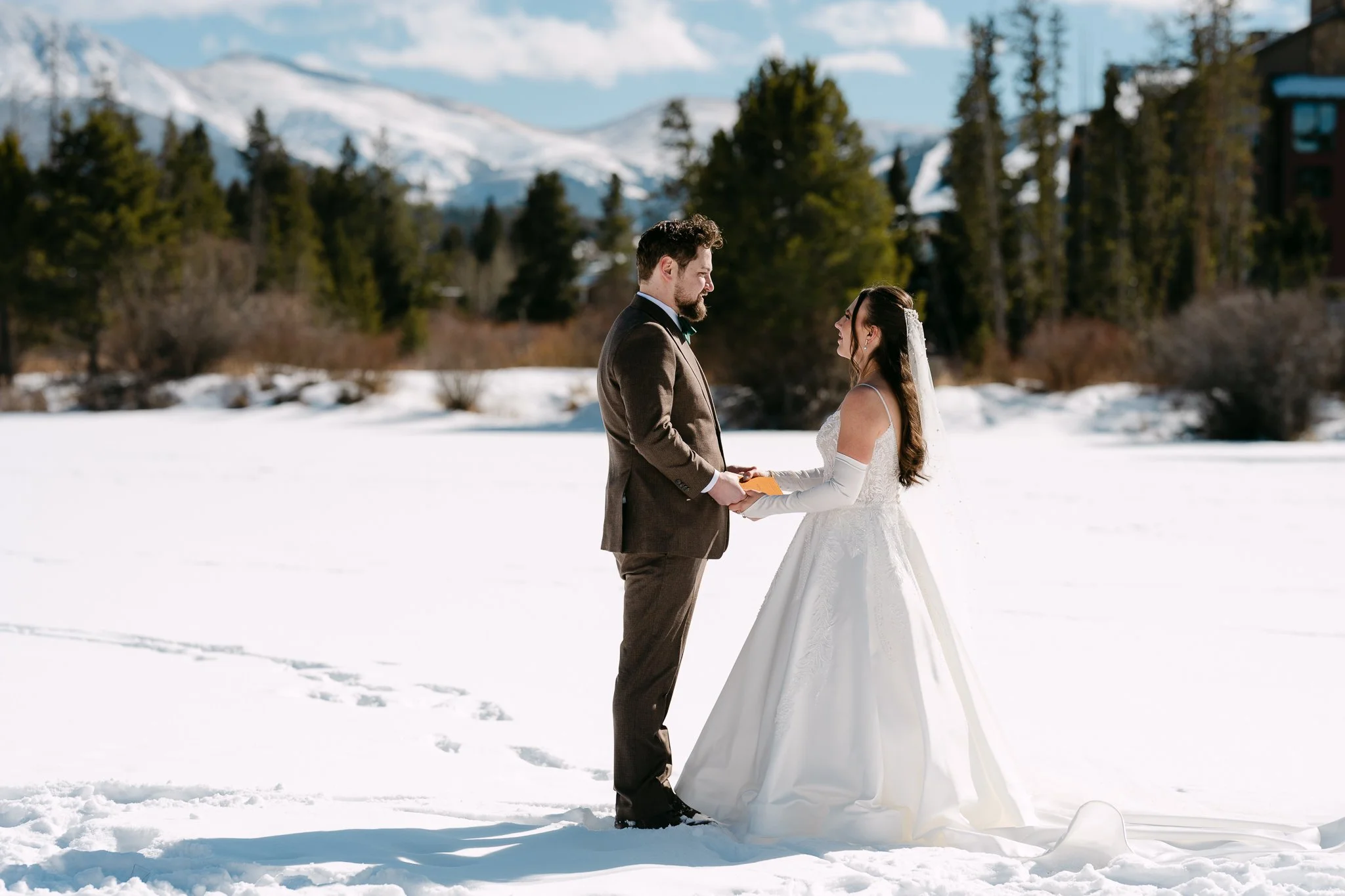 A bride and groom standing in snow, holding hands, outdoors with snow-covered mountains and trees in the background on a sunny day.