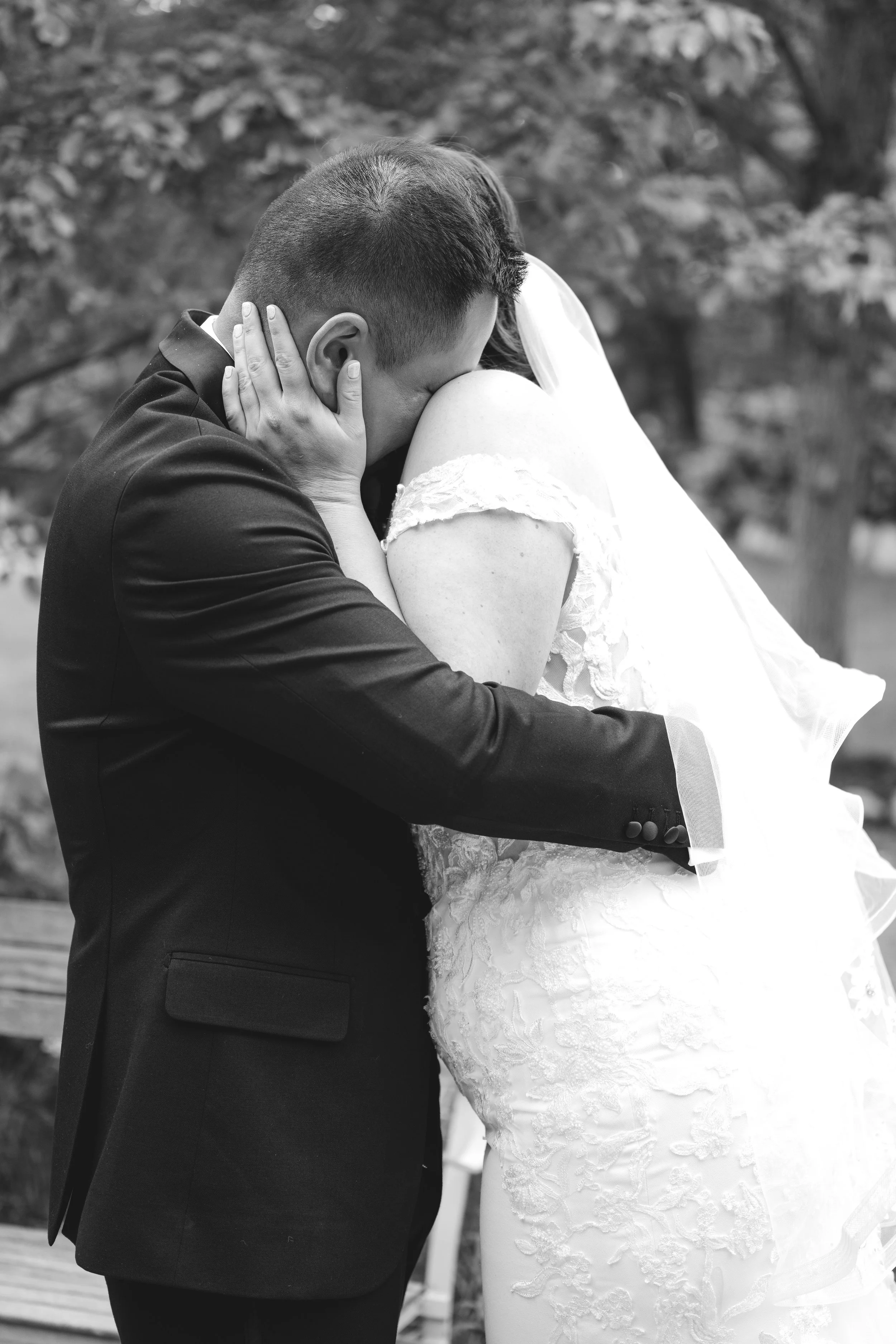 A black and white photo of a couple embracing outdoors, with the woman wearing a white lace dress and veil, and the man in a suit, both holding each other closely.