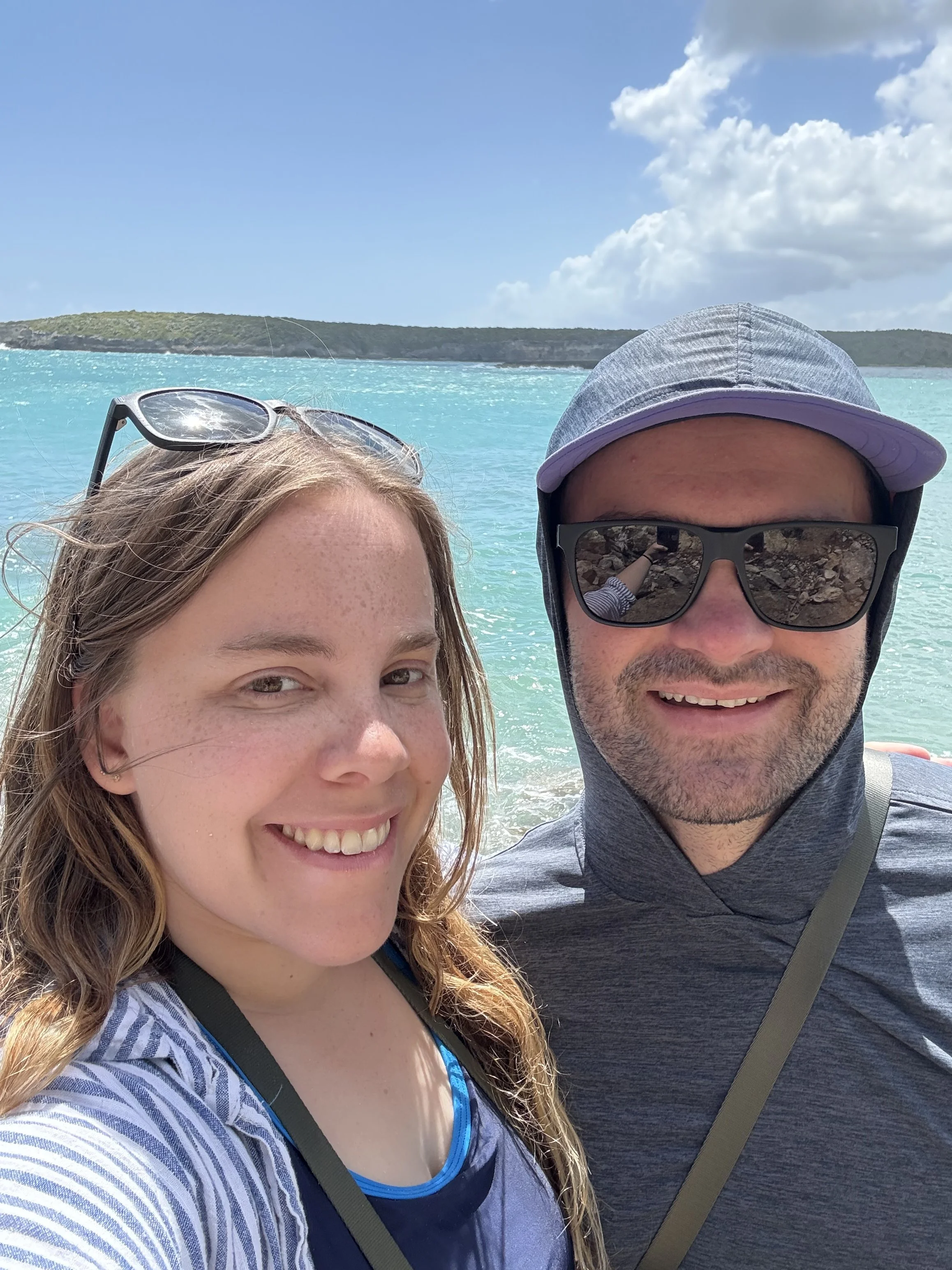 A selfie of a woman and a man on a beach with turquoise water, a distant island, and a partly cloudy sky in the background. The woman has sunglasses on her head, and the man is wearing sunglasses and a hooded shirt.
