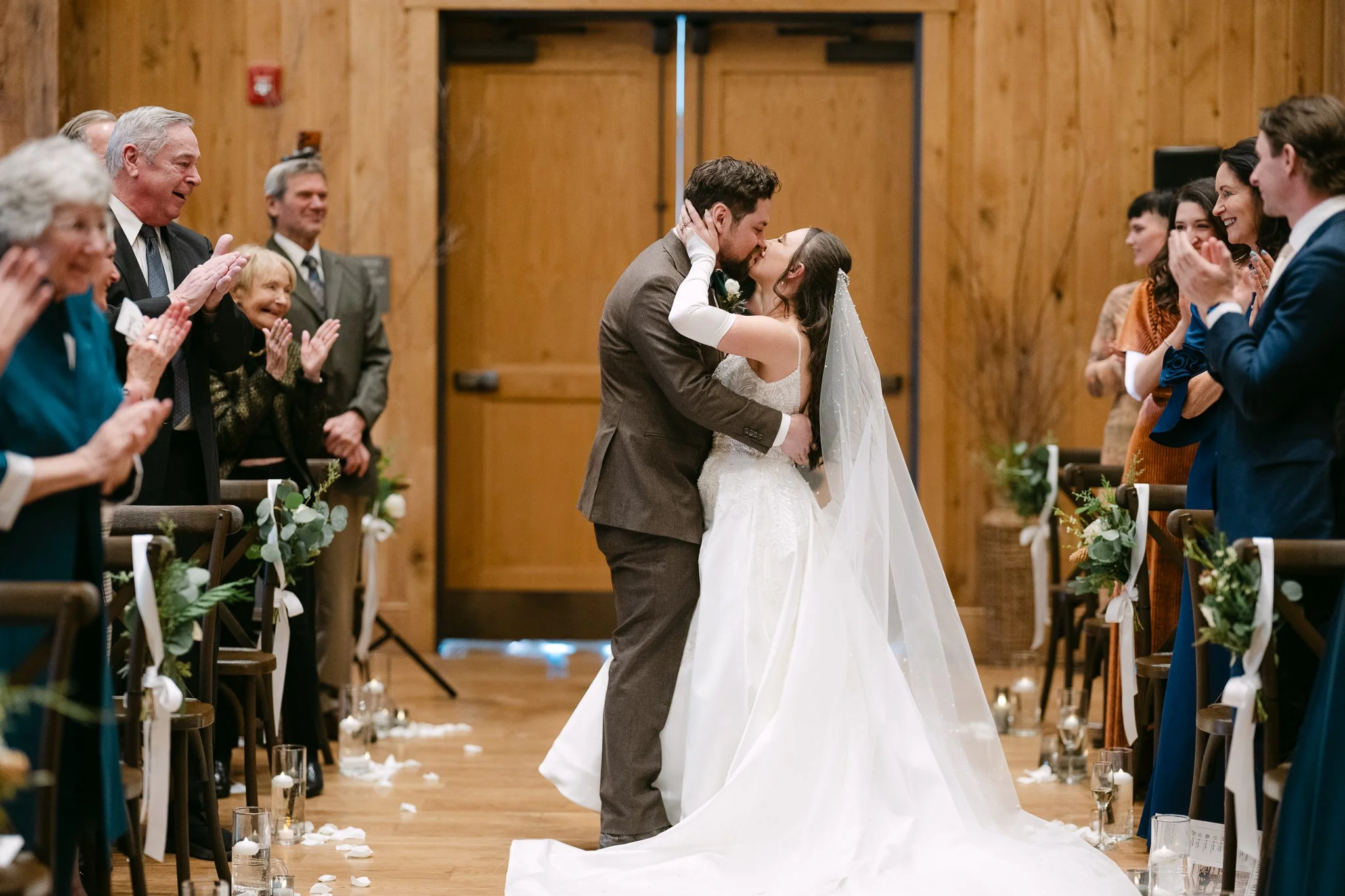 A bride and groom sharing a kiss during their wedding ceremony, surrounded by clapping family and friends in a wooden venue.
