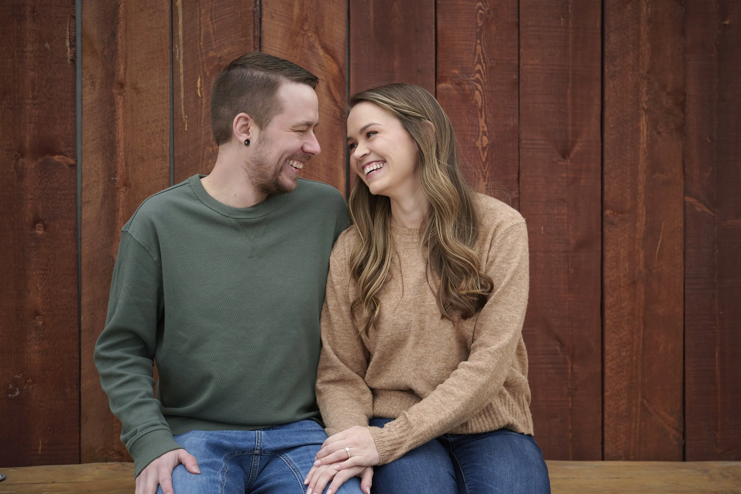 A happy young couple sitting close together on a wooden bench in front of a wooden wall, smiling and enjoying each other's company.