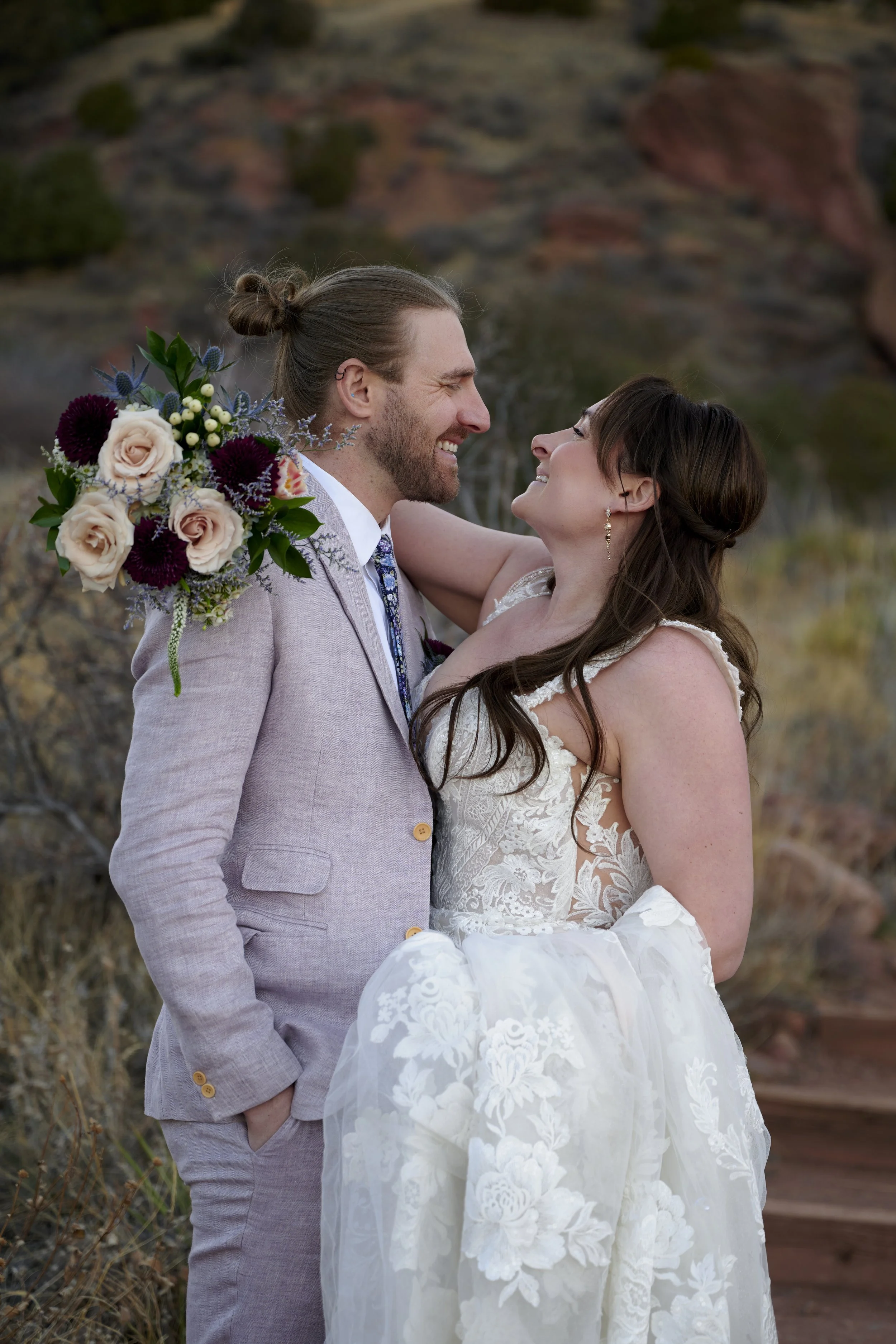 A bride and groom sharing a joyful moment outdoors during their wedding, with the groom holding a bouquet of flowers.