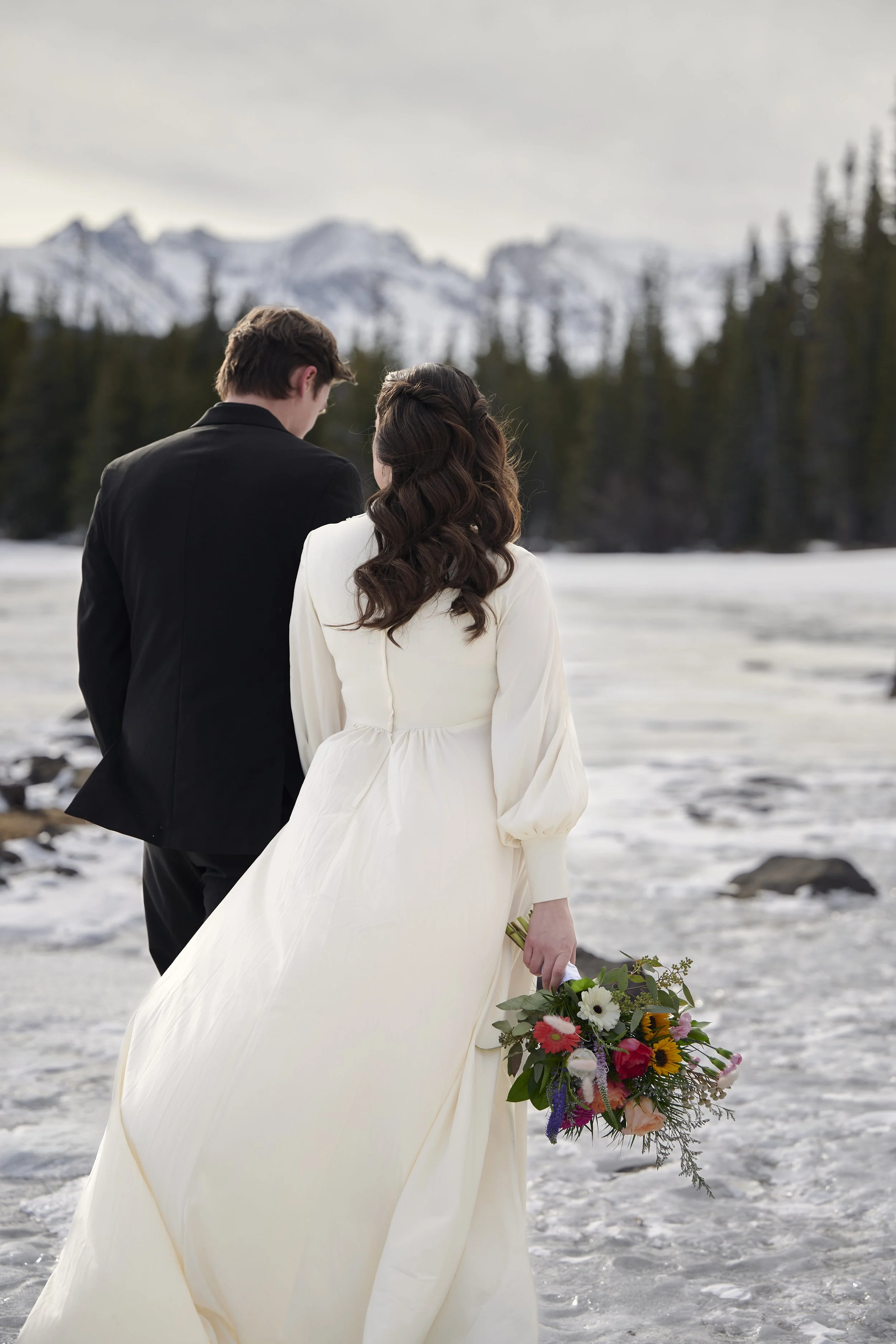A bride and groom standing on ice near a mountain landscape, with the bride holding a bouquet of flowers.