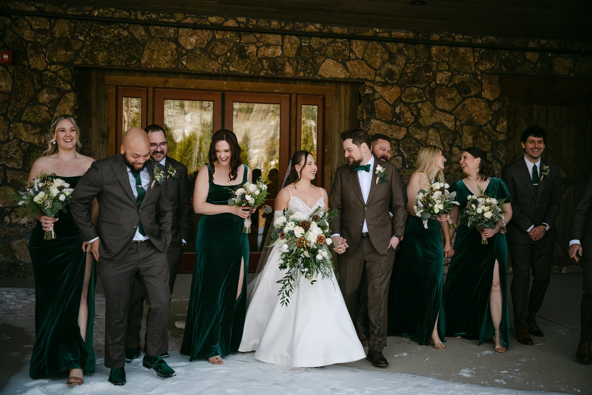 A wedding party standing in front of a stone wall, including the bride in a white dress holding a bouquet of white flowers, the groom in a brown suit, and bridesmaids and groomsmen dressed in dark green and black attire, all smiling and laughing.
