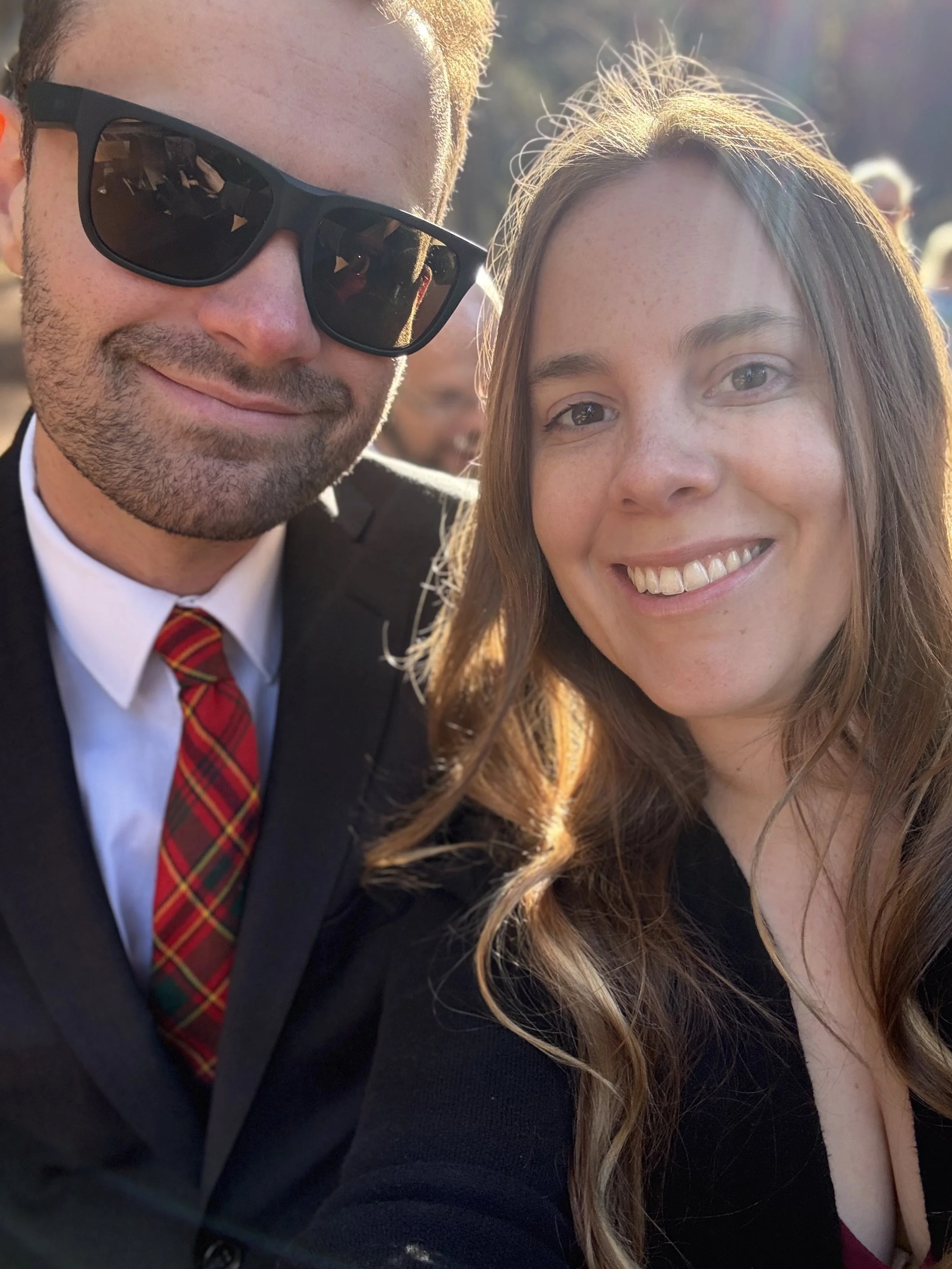 A man and a woman taking a selfie outdoors, smiling, with sunlight behind them.