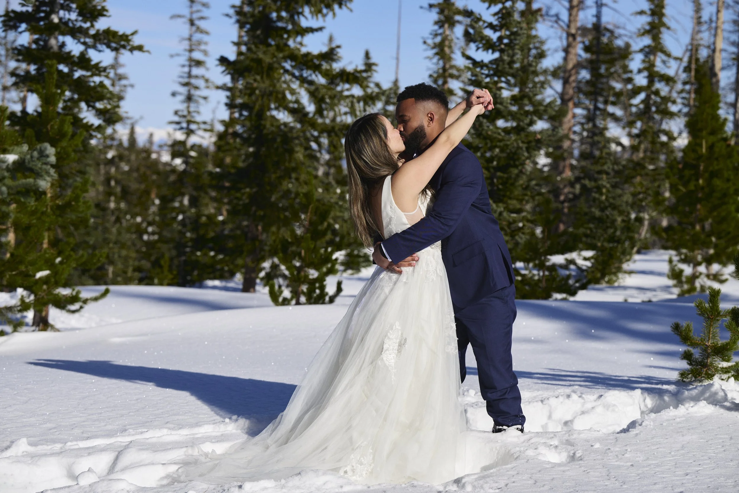 A bride and groom kissing outside in a snowy forest, with the bride in a white wedding dress and the groom in a navy suit.