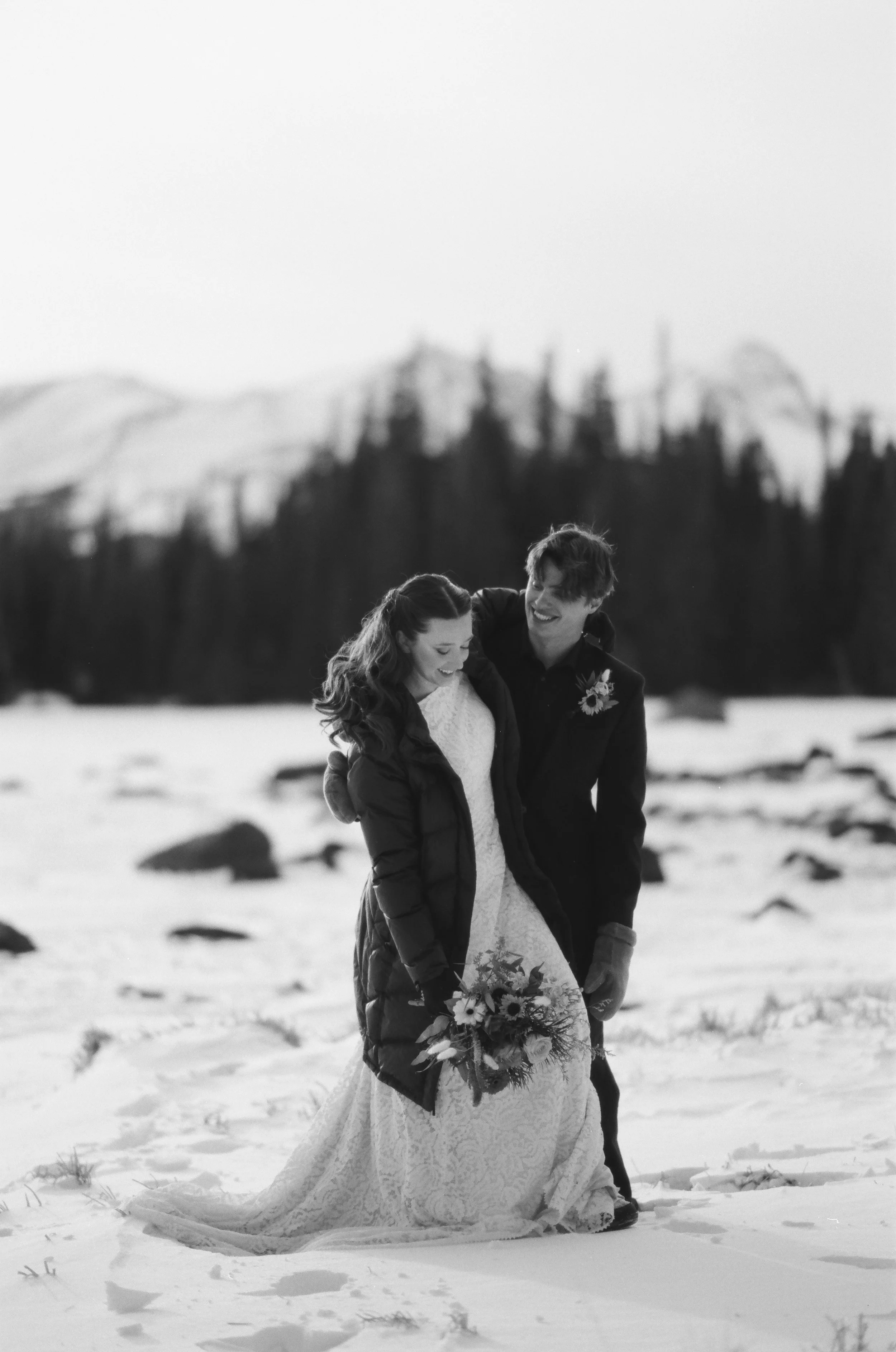 Black and white photo of a couple in wedding attire walking in a snowy landscape, with the bride holding a bouquet, and both smiling.