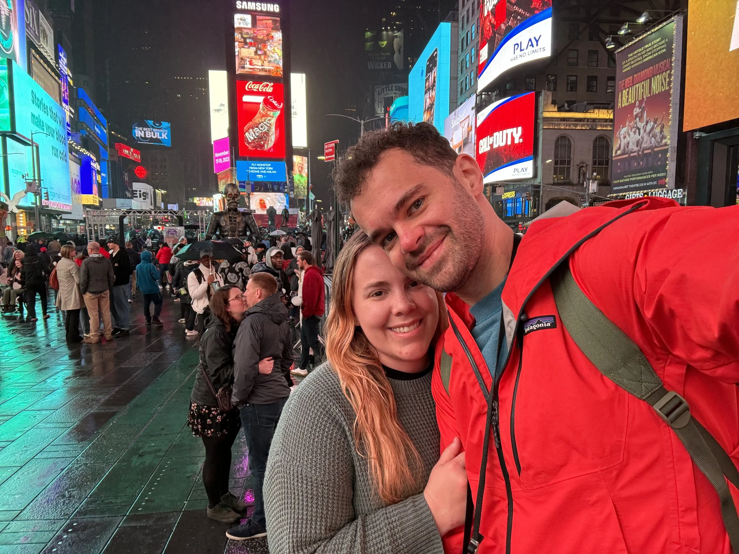 A smiling couple taking a selfie in a busy, brightly lit Times Square at night. The background features numerous digital billboards, advertisements, and a crowd of people, some with umbrellas, on the wet street.