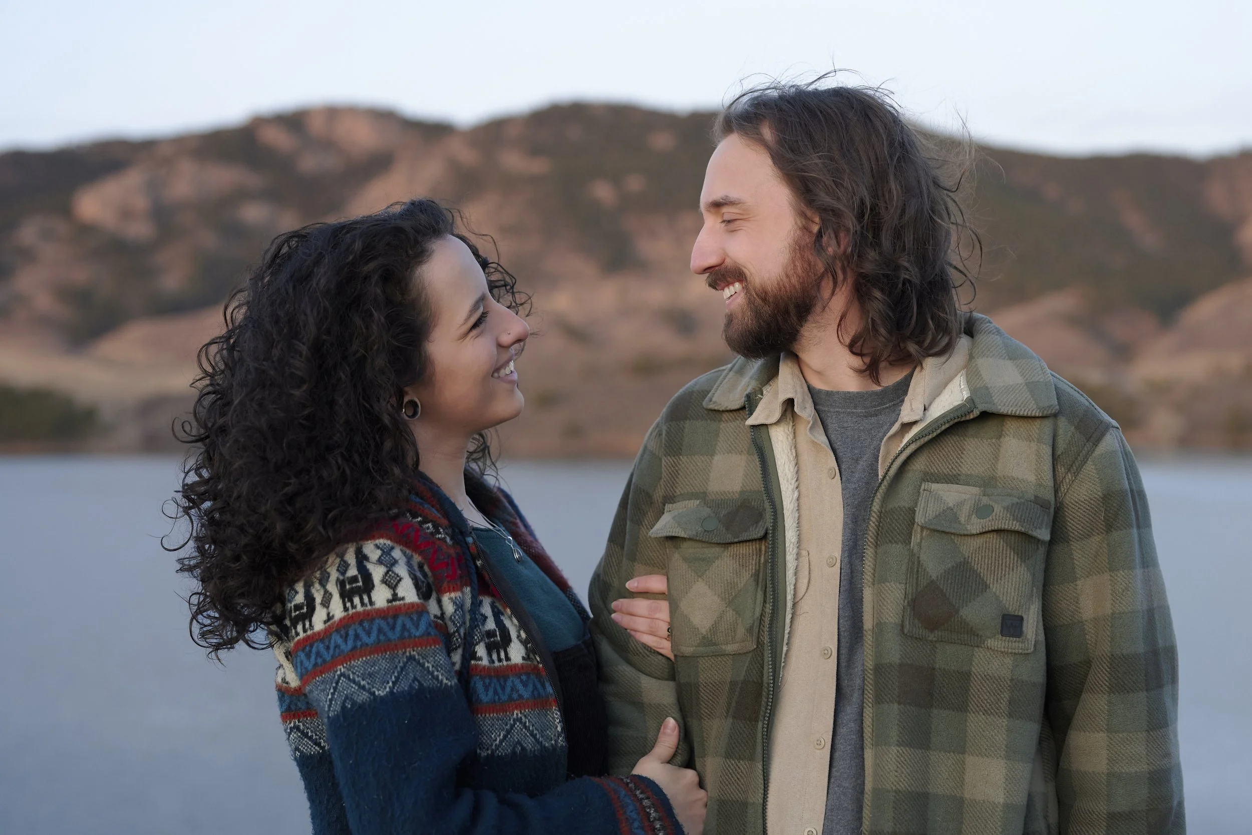 A couple smiling and looking at each other outdoors by a lake, with mountains in the background, during what appears to be late afternoon or early evening.
