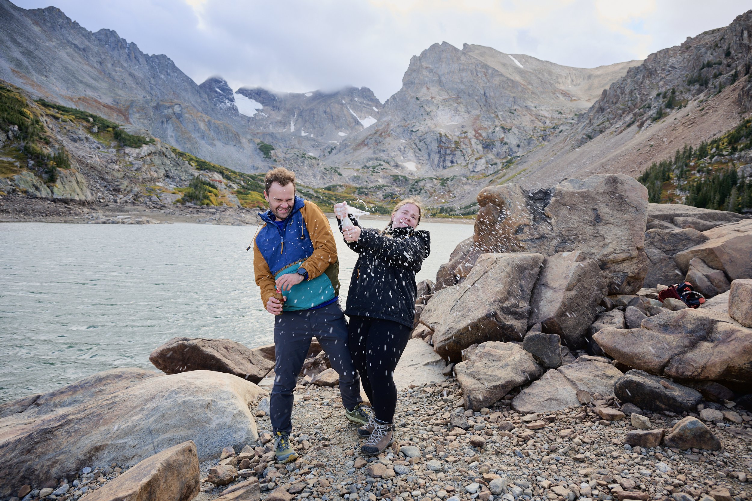 Two people having fun by a mountain lake, one woman squirting water at the man, surrounded by rocky shoreline and mountain peaks in the background.