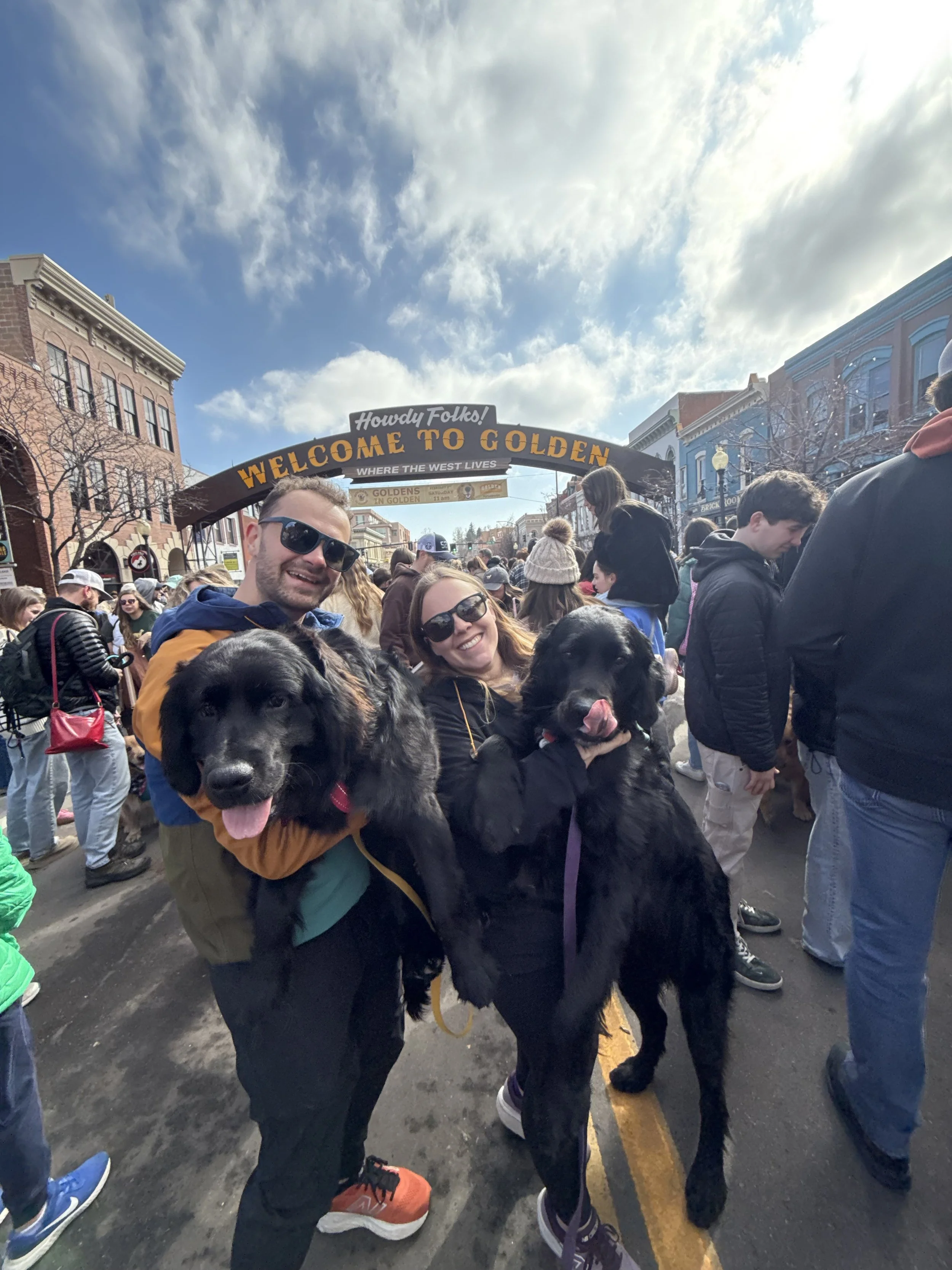 Two smiling people holding large black dogs at a crowded outdoor street event with a welcome sign overhead and colorful buildings in the background.