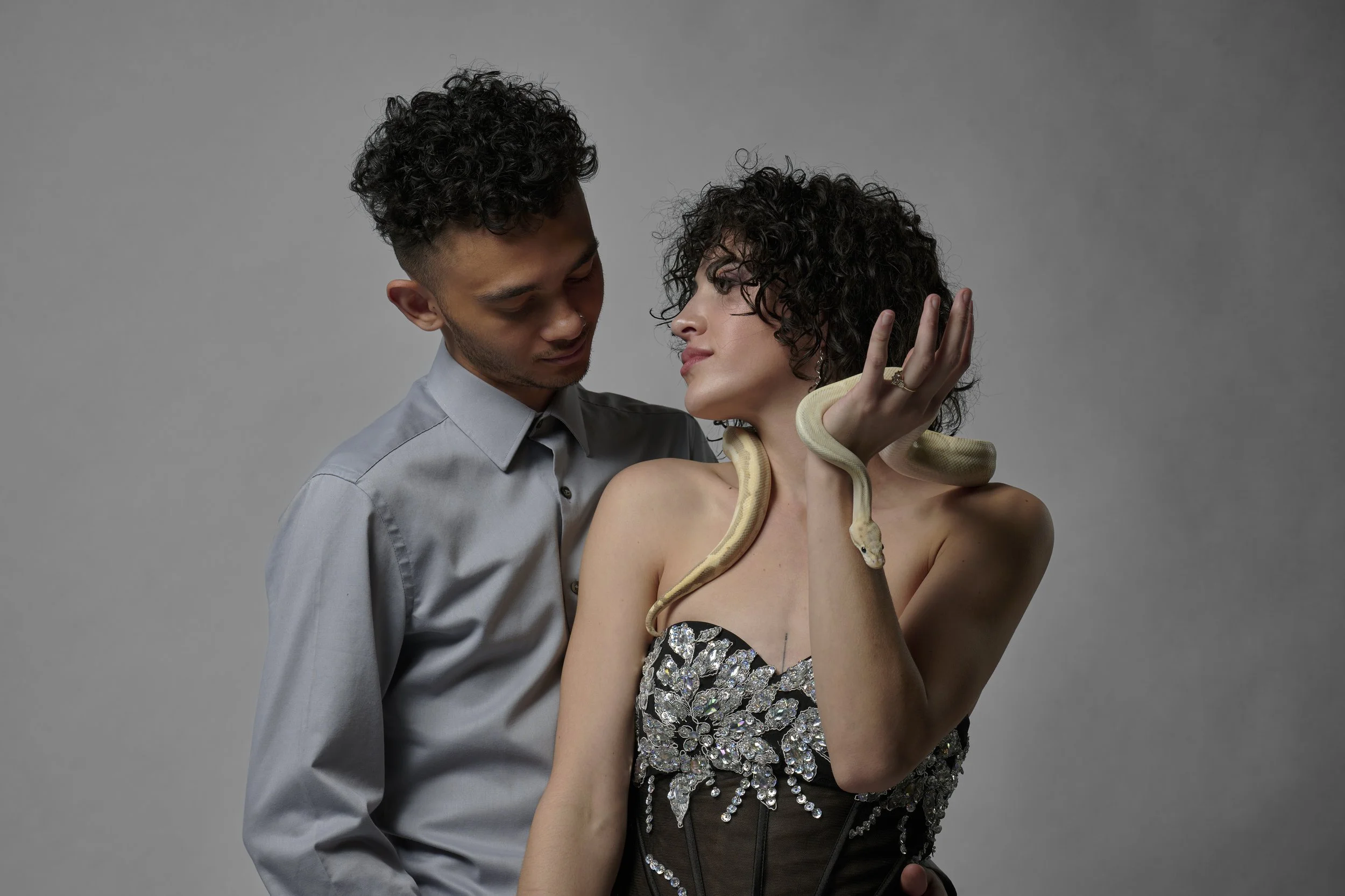A man and woman pose together, with the woman holding a snake around her shoulder and neck. The woman wears a strapless dress with sequins, and the man is in a light gray shirt. They are close, looking into each other's eyes against a gray background