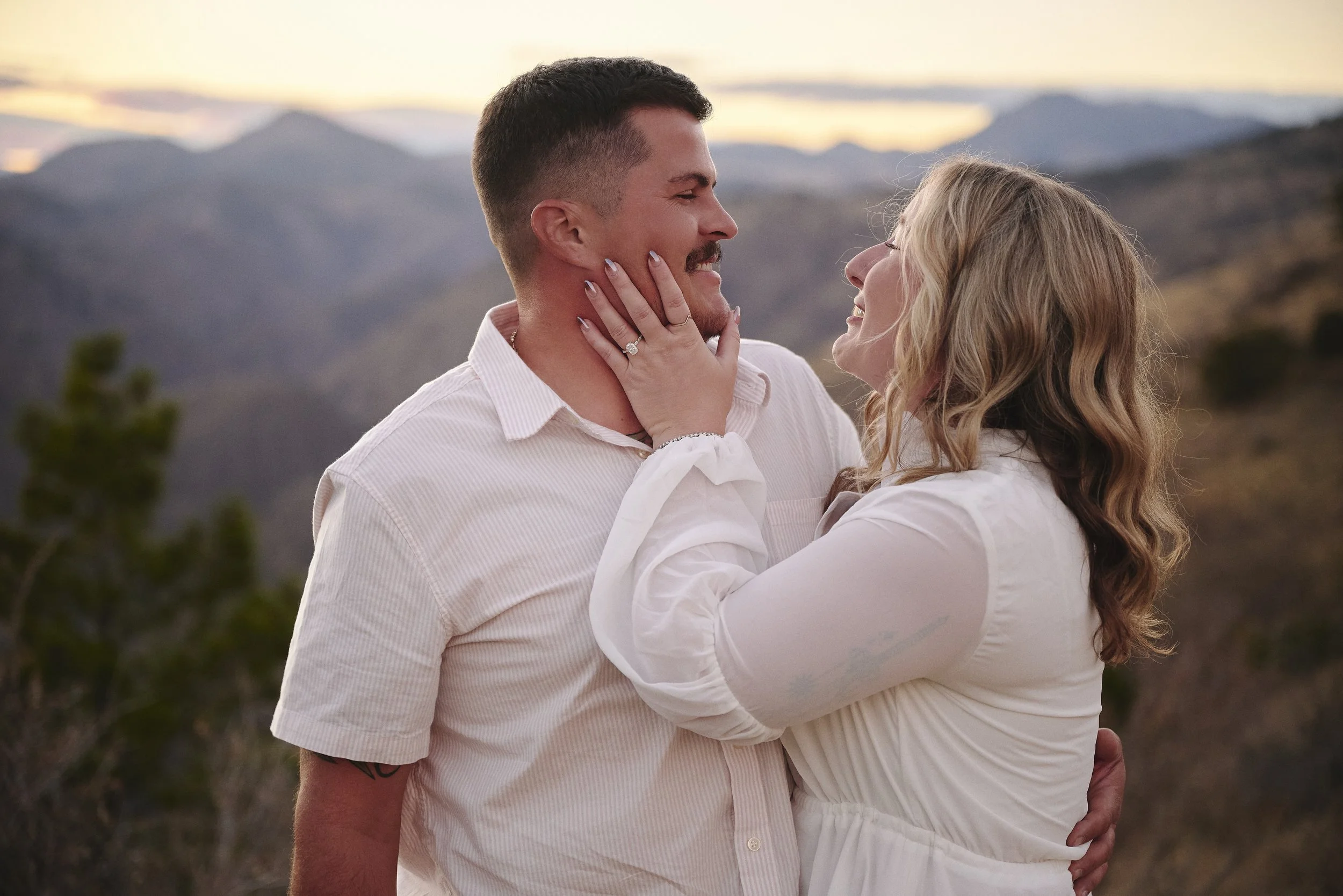 A couple standing outdoors at sunset with mountains in the background, gazing at each other and smiling, with the woman touching the man's face.