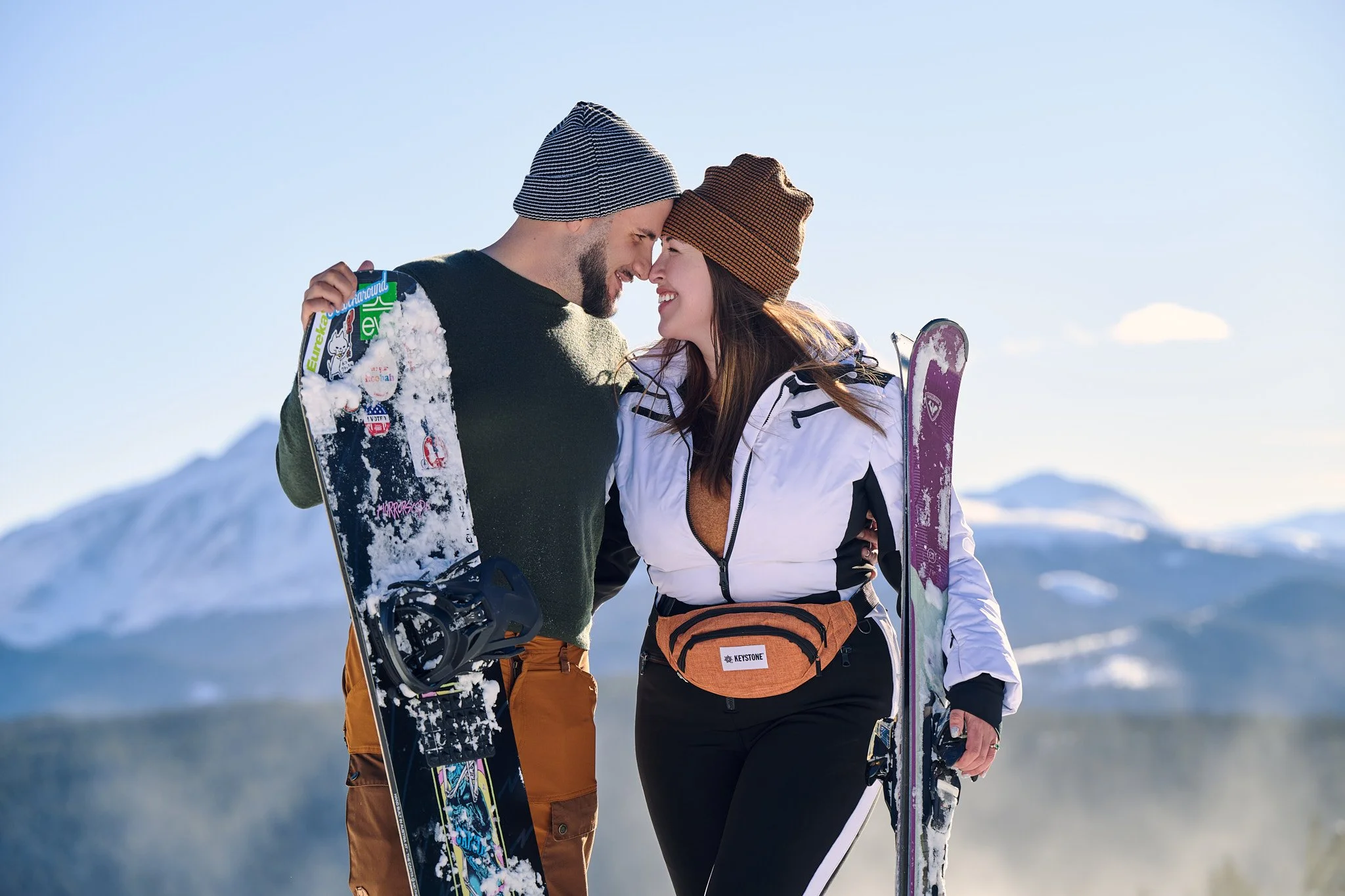 A smiling man and woman dressed in winter sports gear, holding snowboards, standing close together in a snowy mountain landscape with blue sky and distant snow-covered mountains.