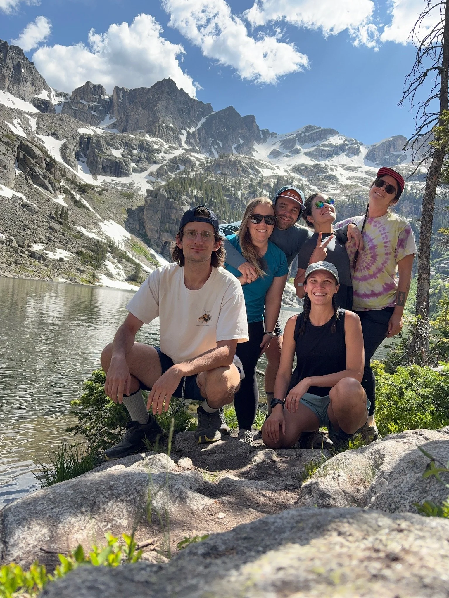 Group of six friends hiking and taking a photo by a mountain lake with snow-capped peaks in the background on a sunny day.