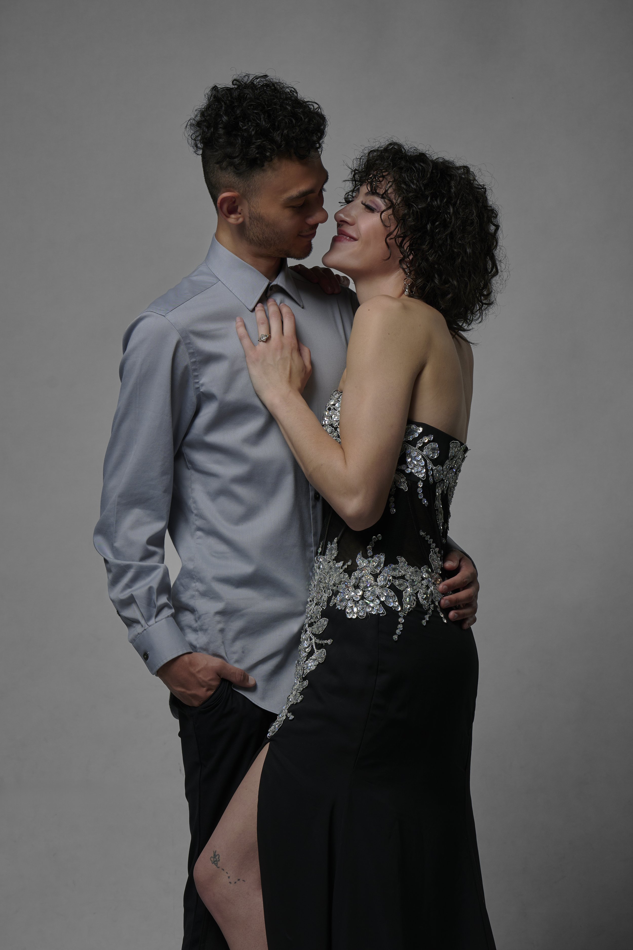 A couple is embracing and looking into each other's eyes against a plain gray background. The woman is wearing a strapless, black dress with silver embellishments and a thigh-high slit. The man is dressed in a light gray, long-sleeved button-up shirt with his hand in his pocket.