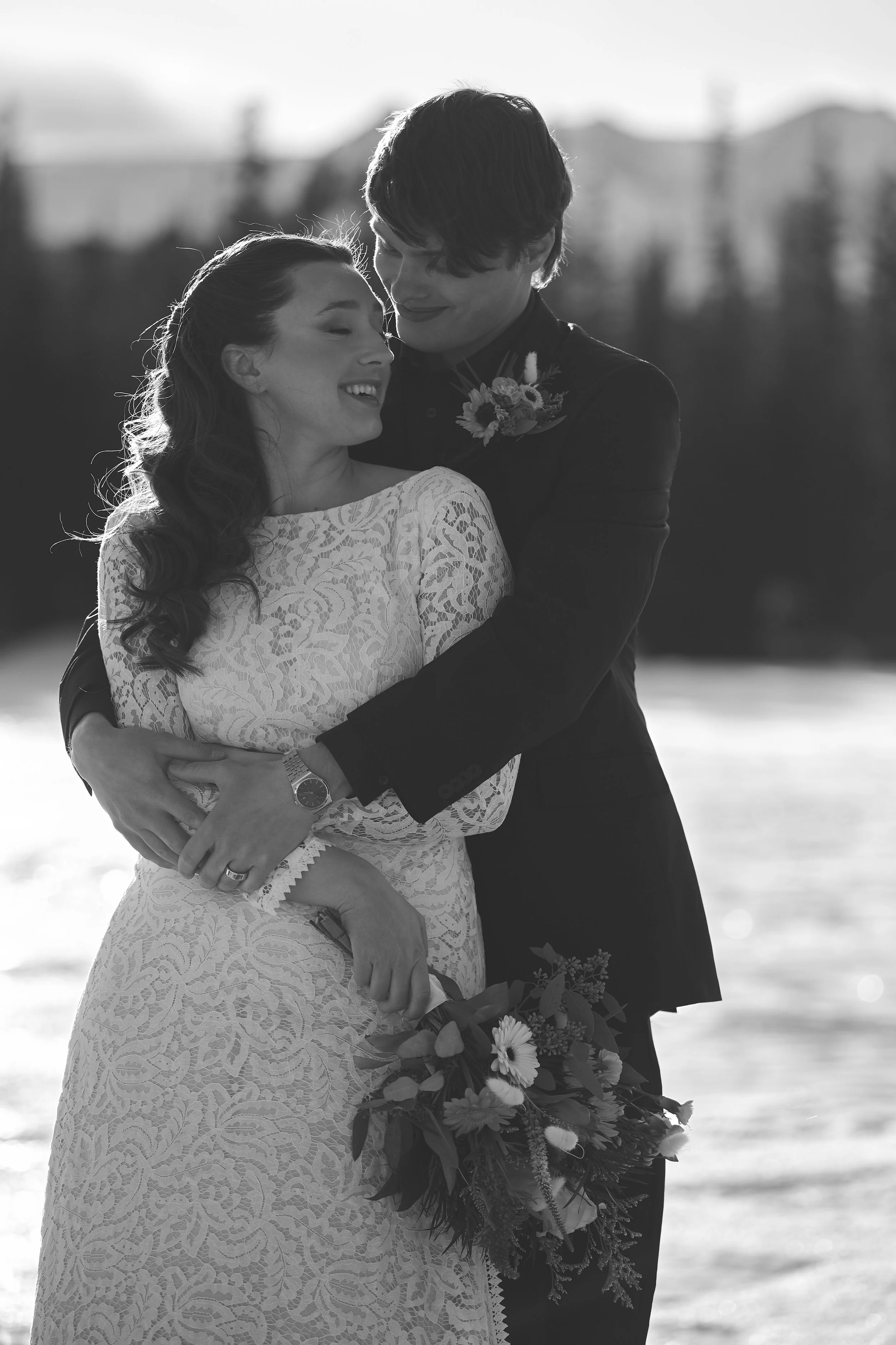 A black and white photo of a joyful couple embracing outdoors during the daytime, with buildings and a blurred background, dressed in wedding attire, and holding a bouquet of flowers.