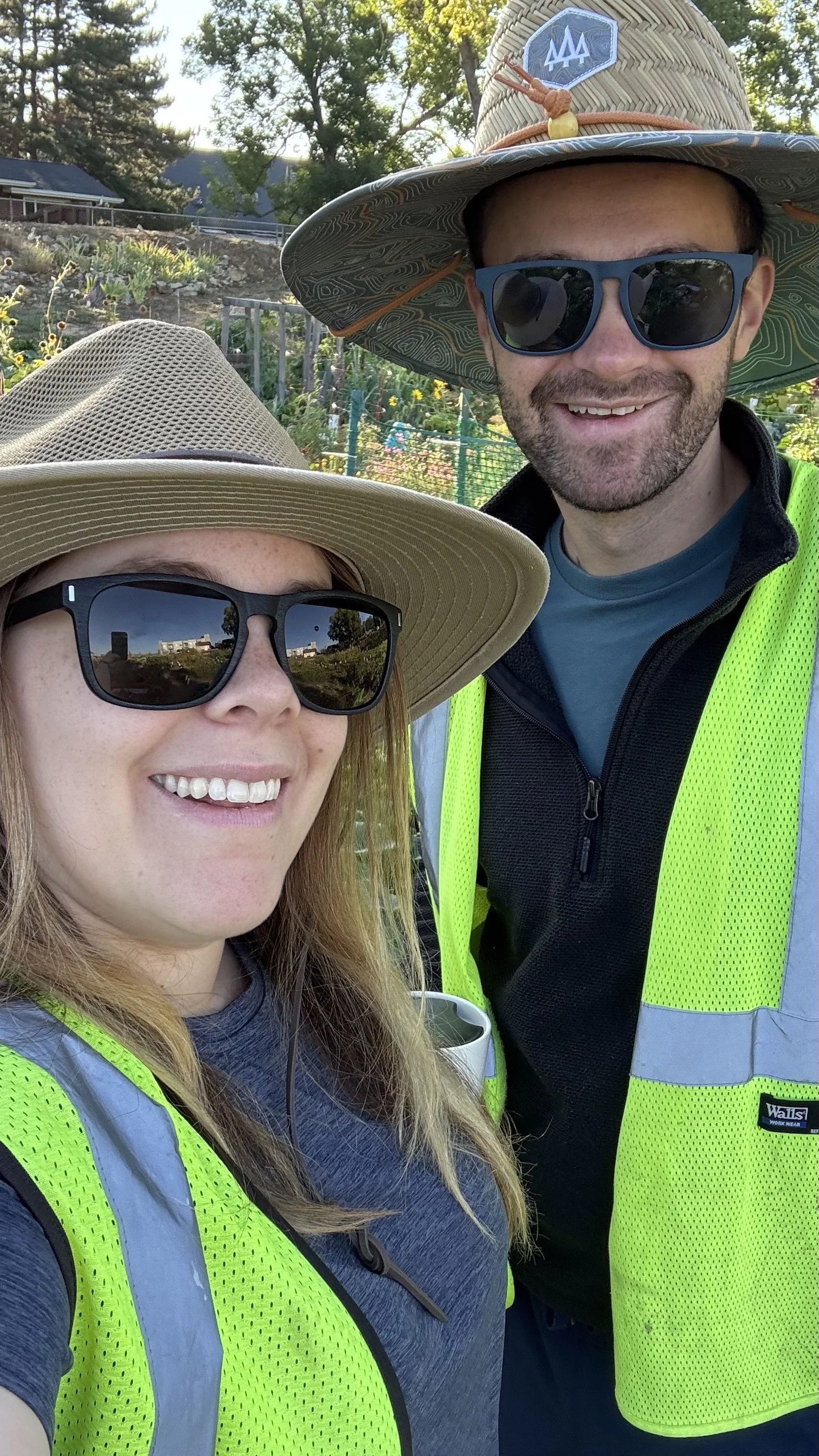 Two smiling people wearing sunglasses and wide-brimmed hats, standing outdoors in a sunny garden or farm, both wearing bright yellow safety vests.