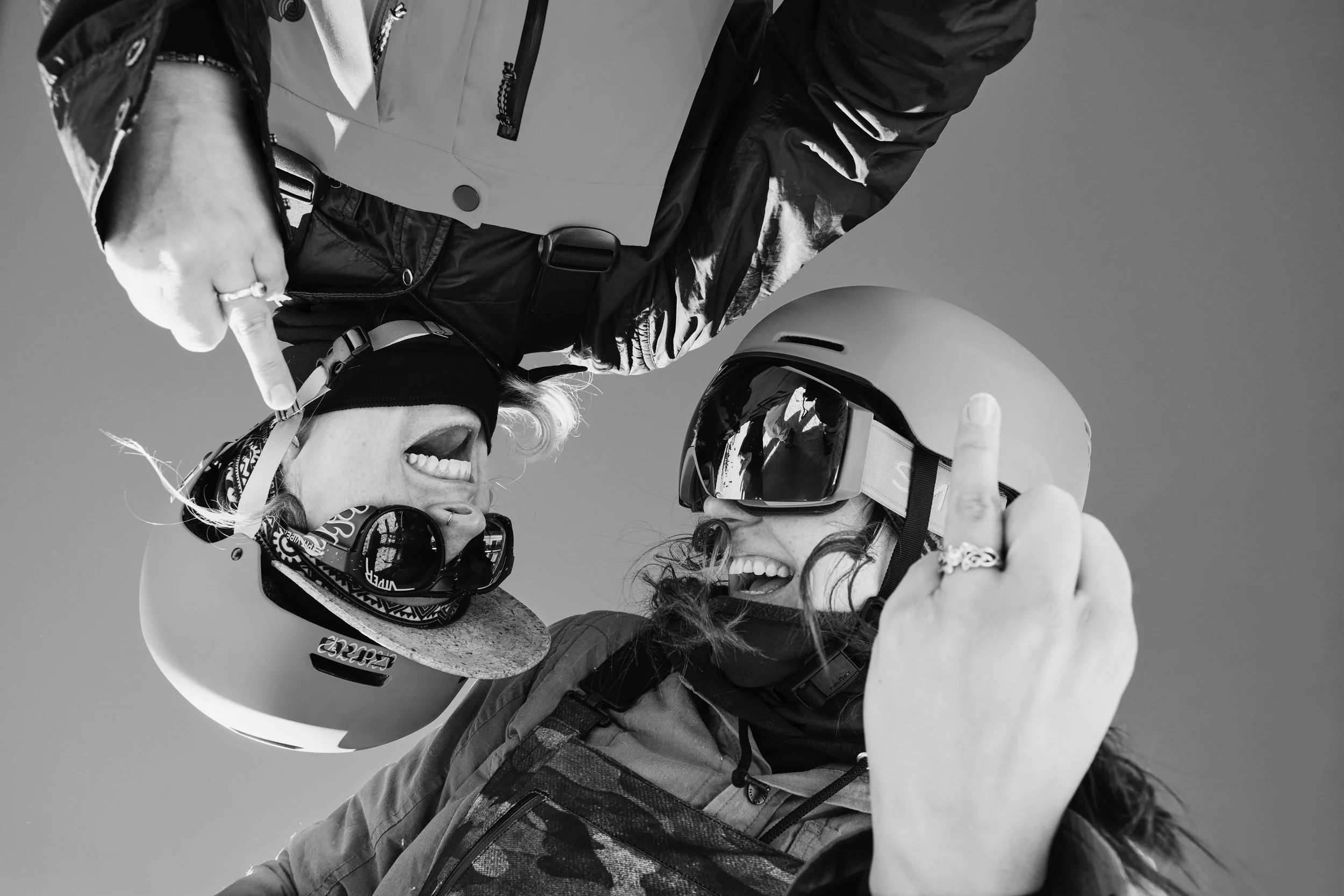 Two women, one wearing a helmet and goggles, and the other wearing goggles and a bandana, are smiling and giving each other a high five against a clear sky.