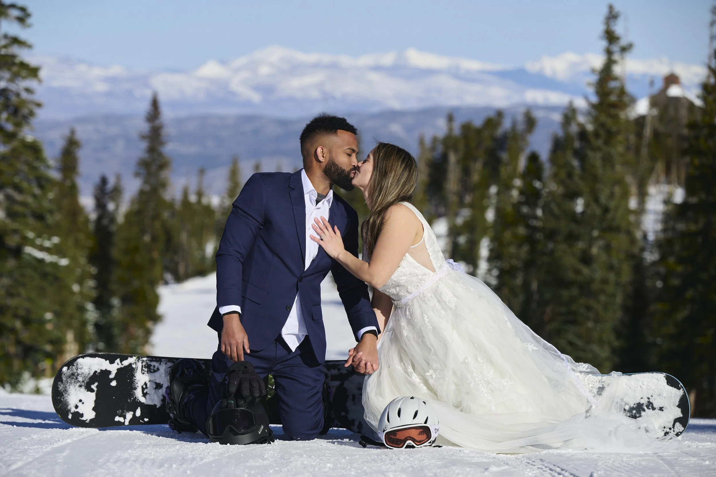 A newlywed couple kissing on snow with snowboards, wearing wedding attire, against a mountain and forest background.