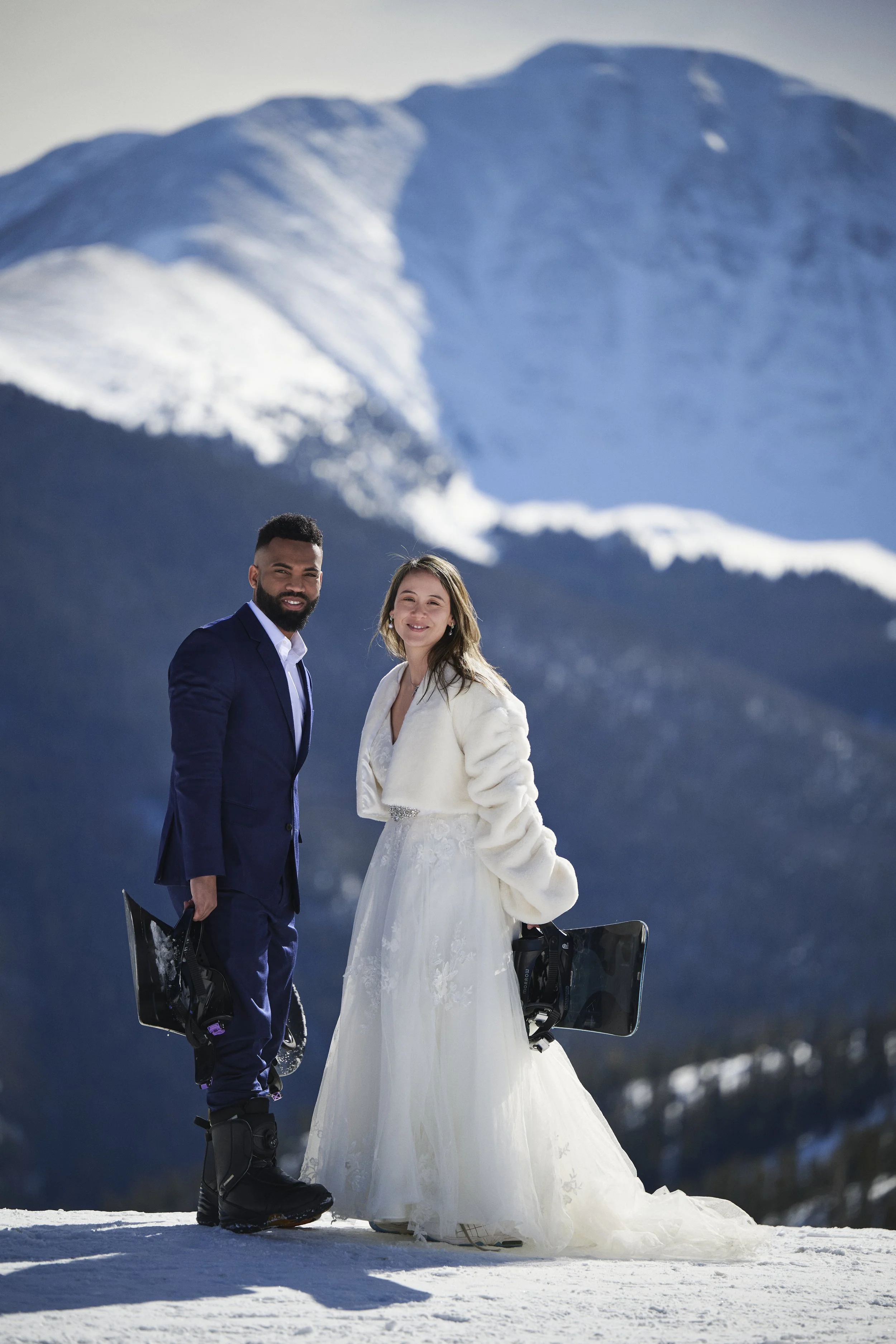 A bride and groom standing on snow-covered ground with mountainous snow-capped peaks in the background. The groom is in a navy blue suit holding a snowboard, and the bride is in a white wedding dress with a white coat, also holding a snowboard.