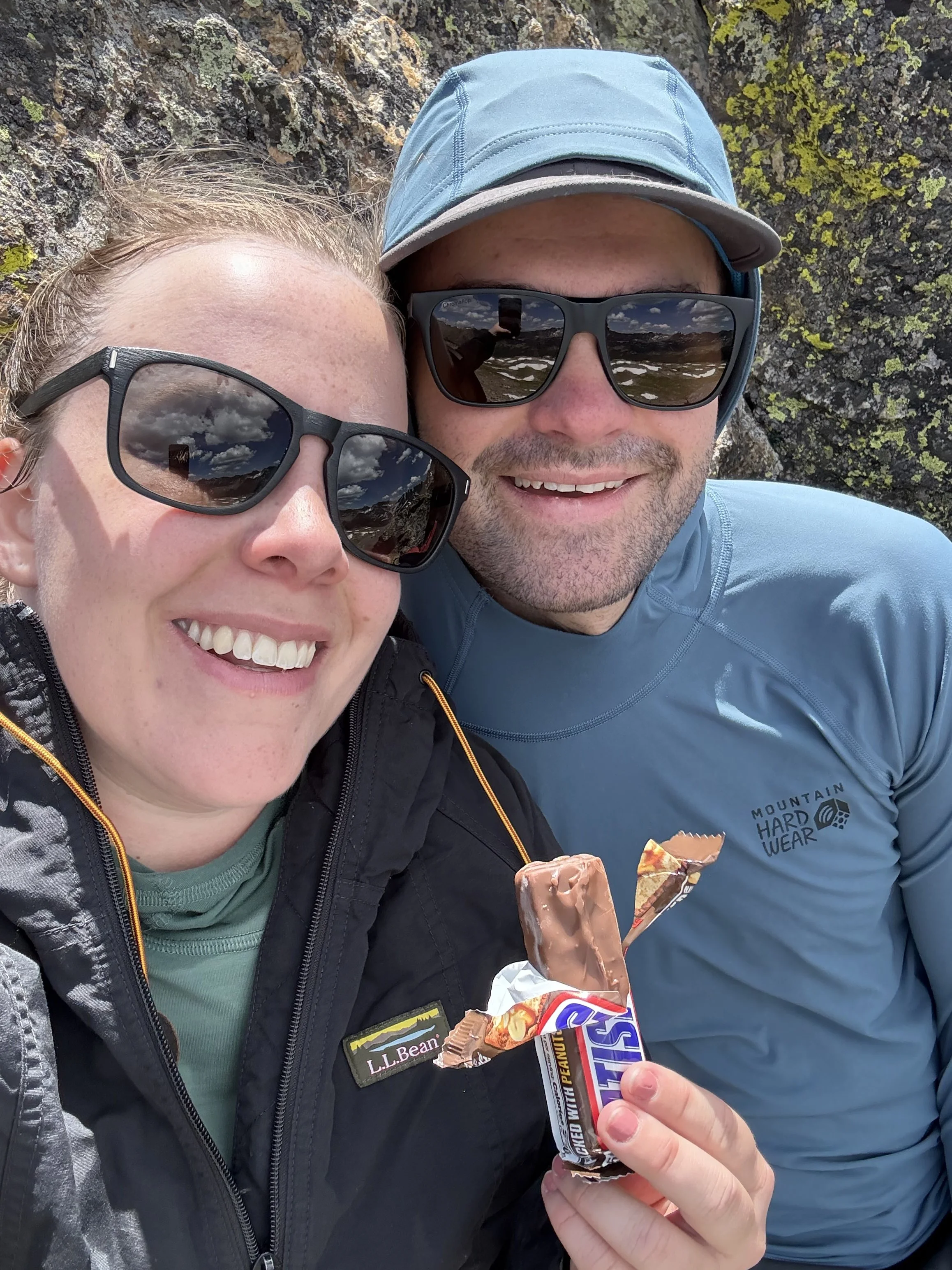 A smiling woman and man outdoors, both wearing sunglasses, with a rocky background. The woman is holding a partially unwrapped Snickers peanut butter bar.