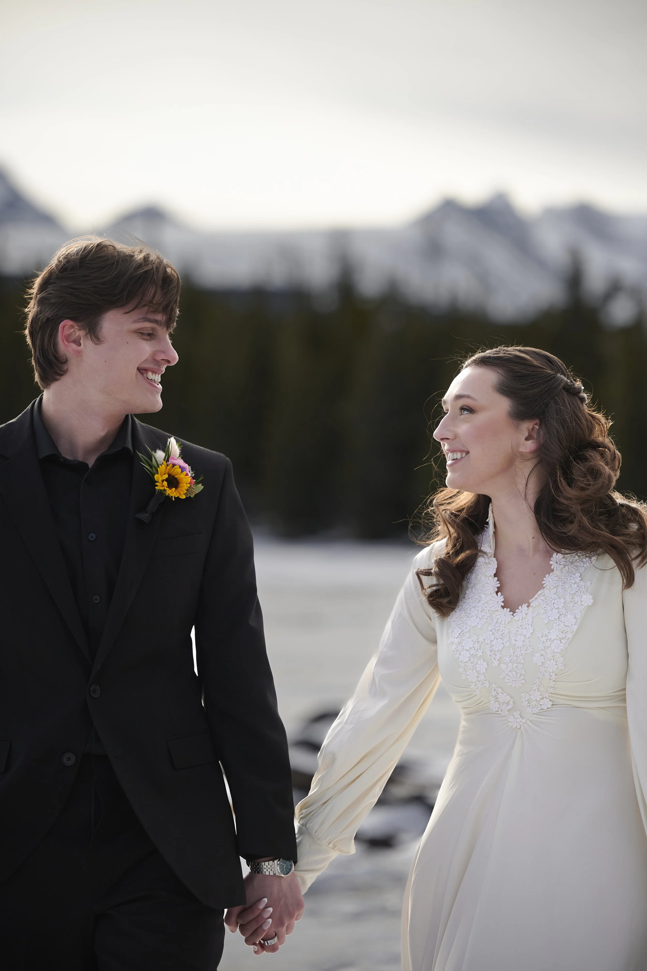 A couple holding hands and smiling at each other outdoors in winter, with snow-covered mountains in the background.