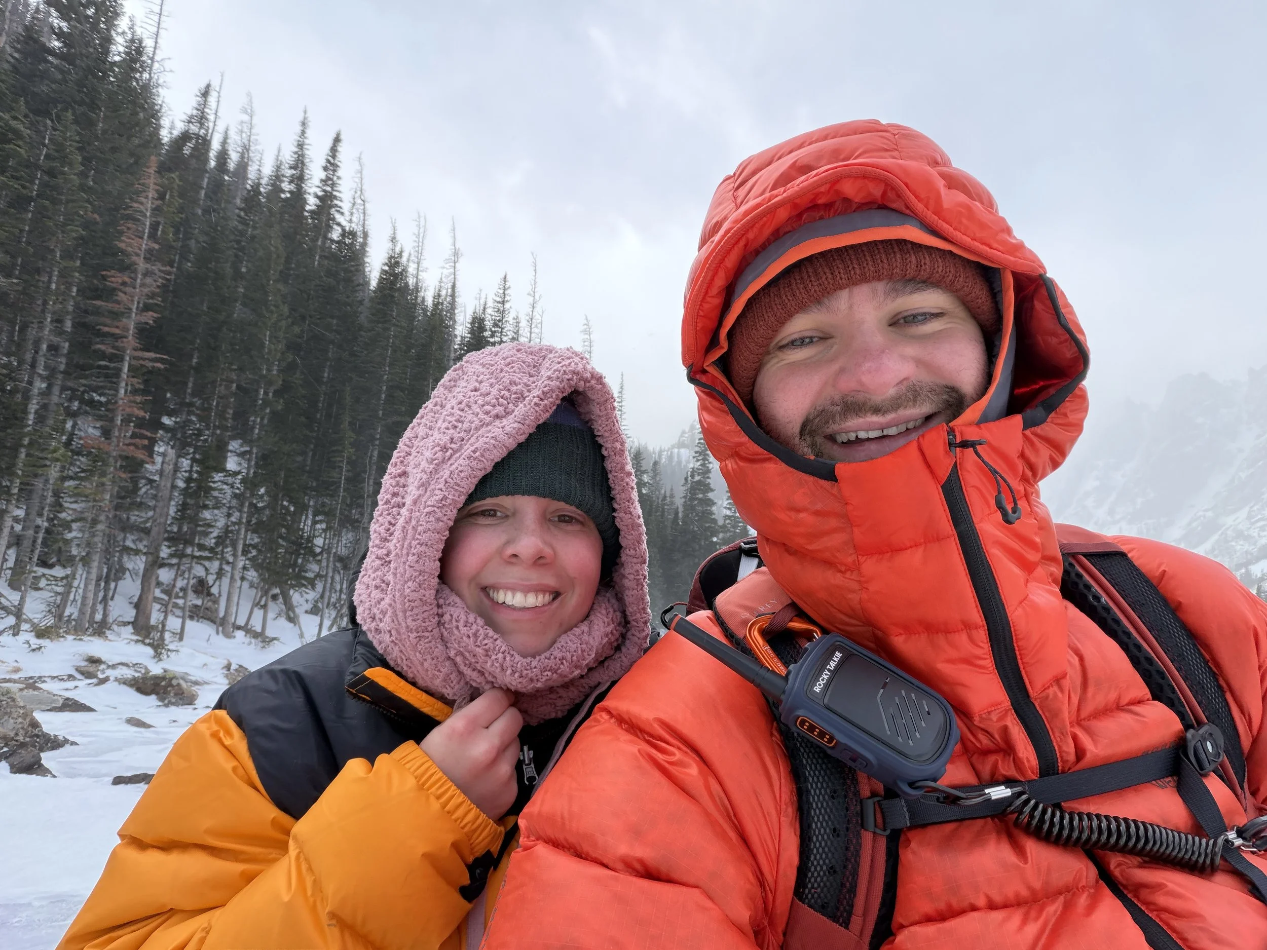 Two people in winter gear take a selfie outdoors in a snowy mountainous landscape with trees and fog in the background.