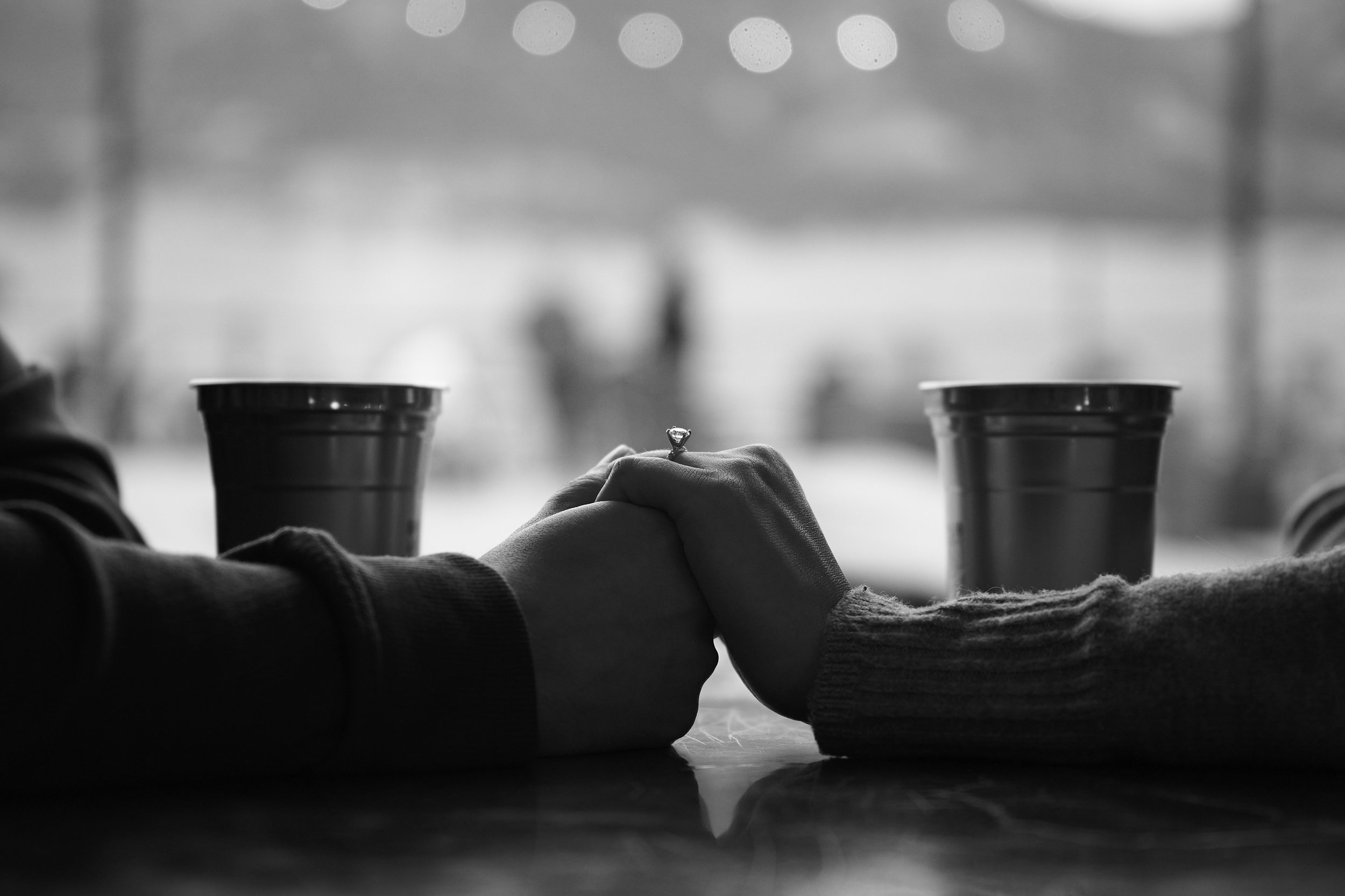 Black and white photo of two people holding hands with a ring on one person's finger, sitting at a table with two cups, with a blurred background of a window and outdoors.