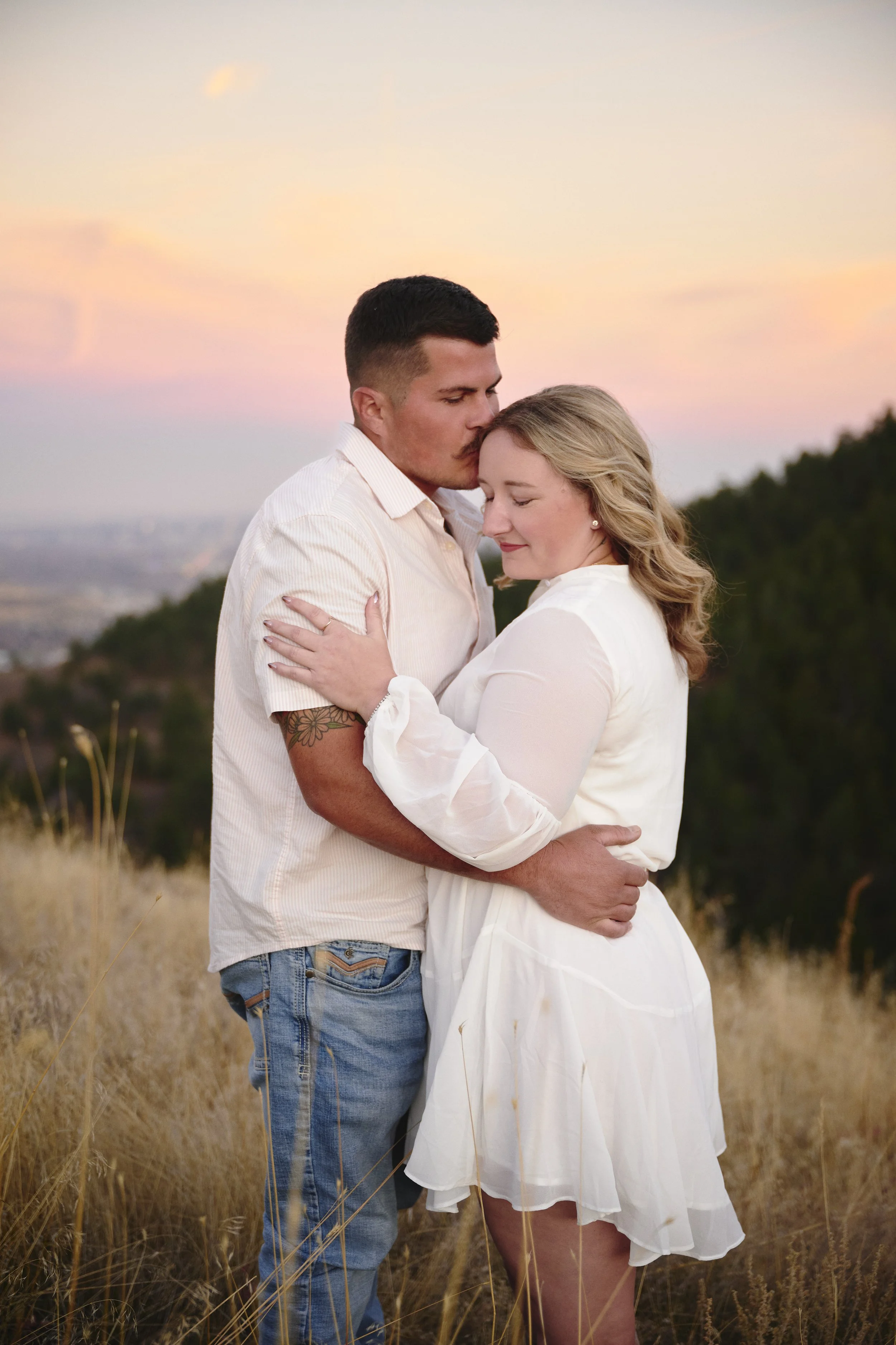 A couple embracing outdoors during sunset, with pink and orange skies and grassy hills in the background.