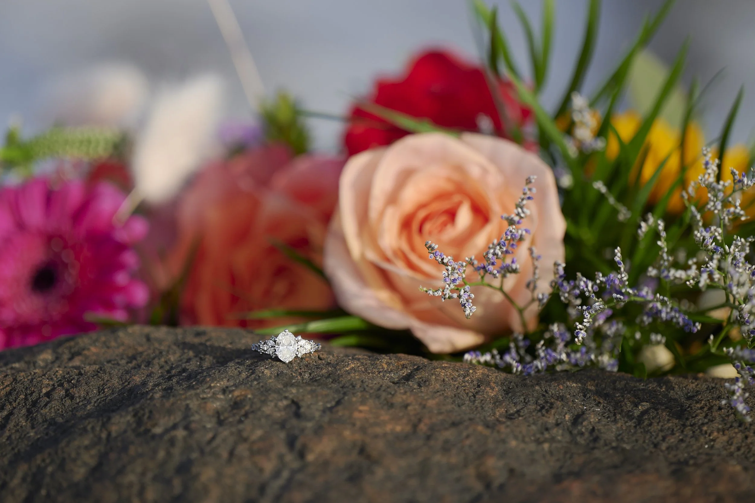 A diamond ring placed on a textured dark stone surface with a bouquet of pink, red, peach, yellow, purple, and white flowers in the background."}