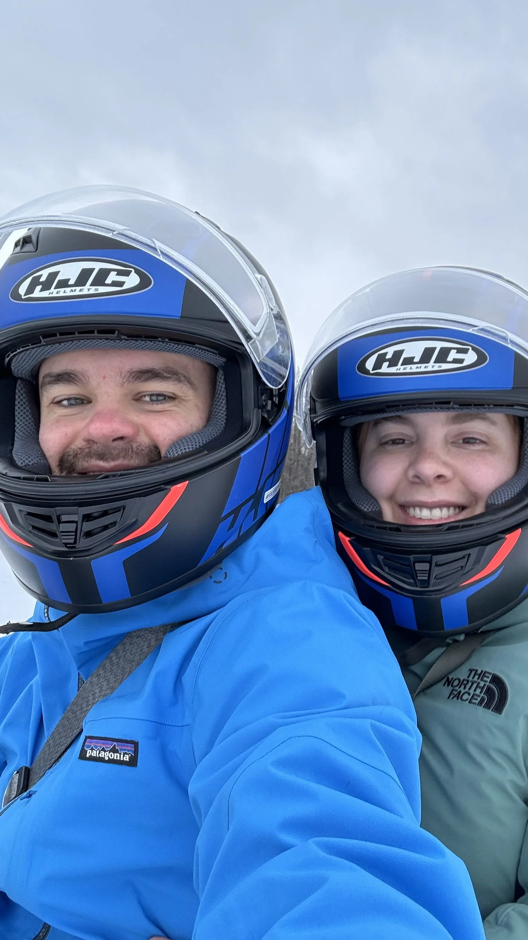 Two people in blue and green jackets wearing black and blue HJC motorcycle helmets taking a selfie outdoors.