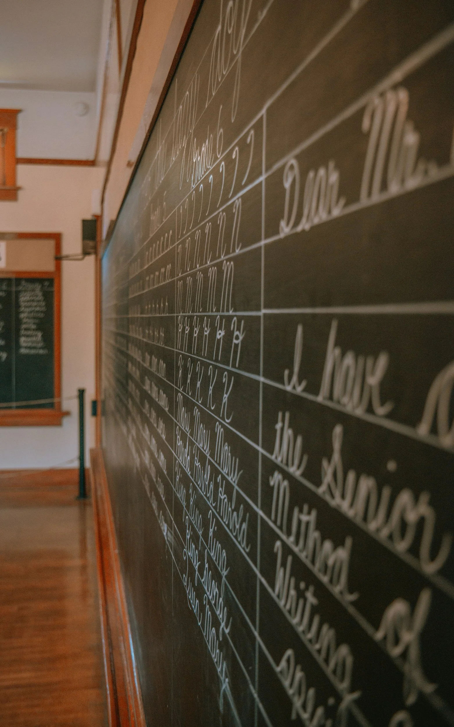 Close-up of a classroom chalkboard with handwritten schedule or lessons, seen at an angle from the right, with a wooden floor and partial view of an adjacent blackboard in the background.