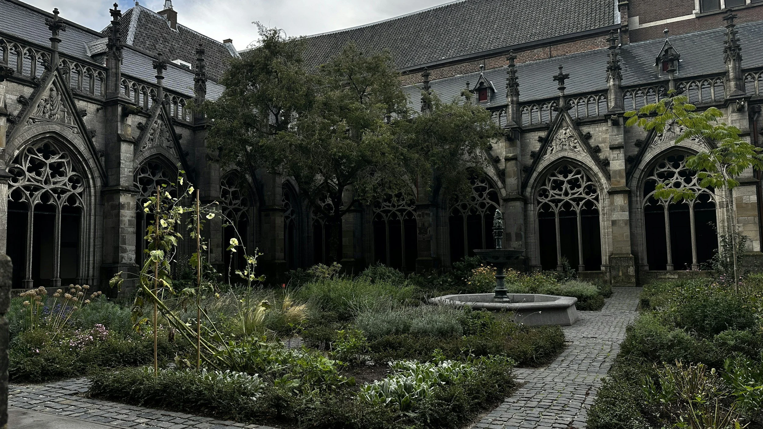 A historic stone church with gothic windows and intricate architecture surrounds a small garden with a pathway, a fountain, and various plants.