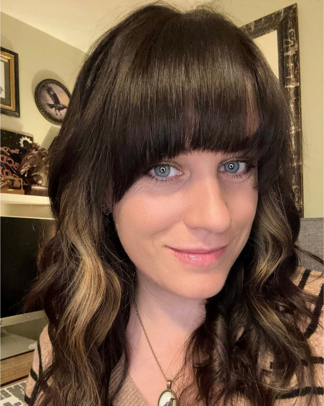 Close-up of C.H.Folan,  a woman with dark brown curly hair, straight bangs, and blue eyes, smiling softly, wearing a necklace with an oval pendant, in a room with artwork and figurines in the background.