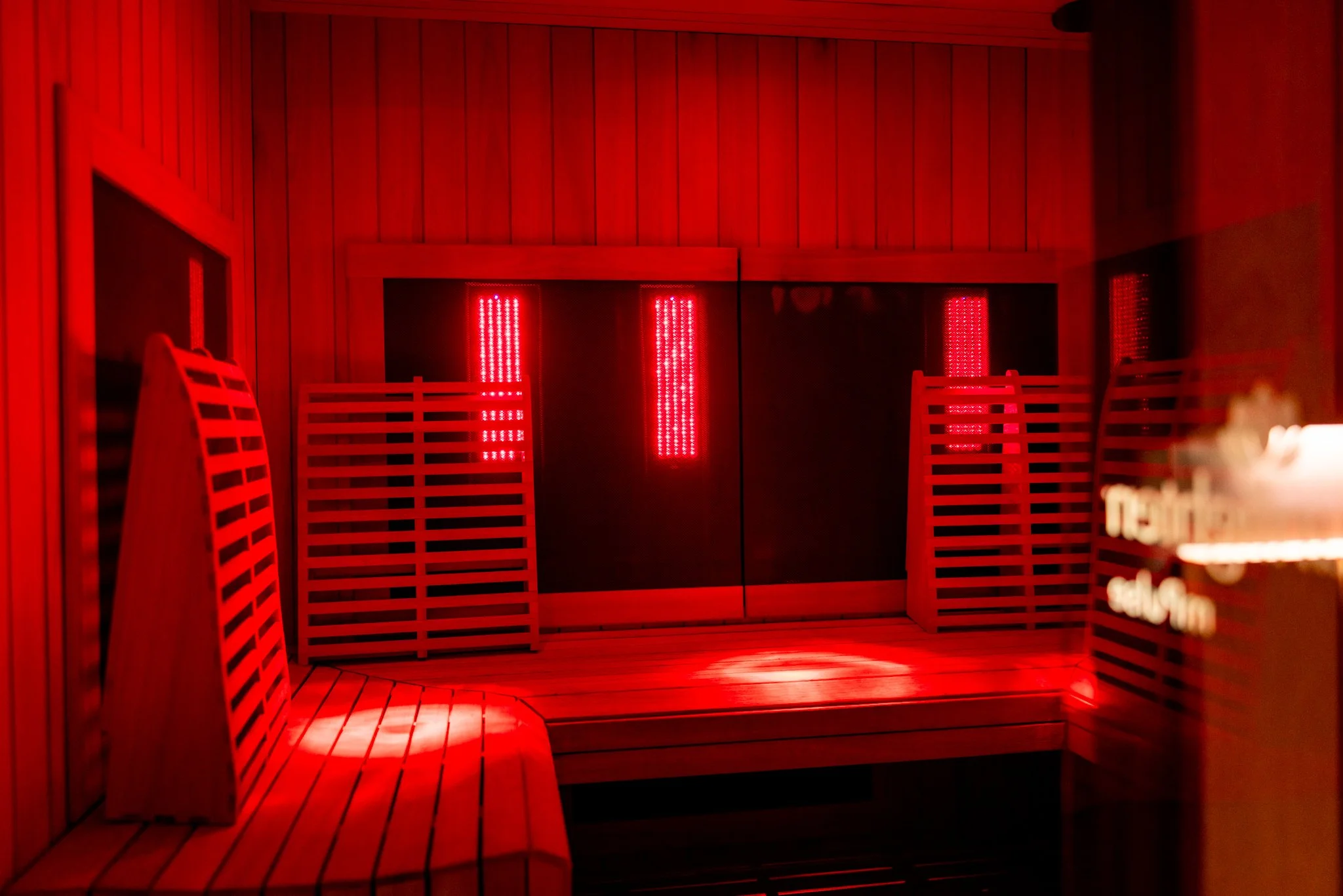 Inside a large wooden infrared sauna room illuminated by red light panels with cedar wooden benches and slatted backrests.