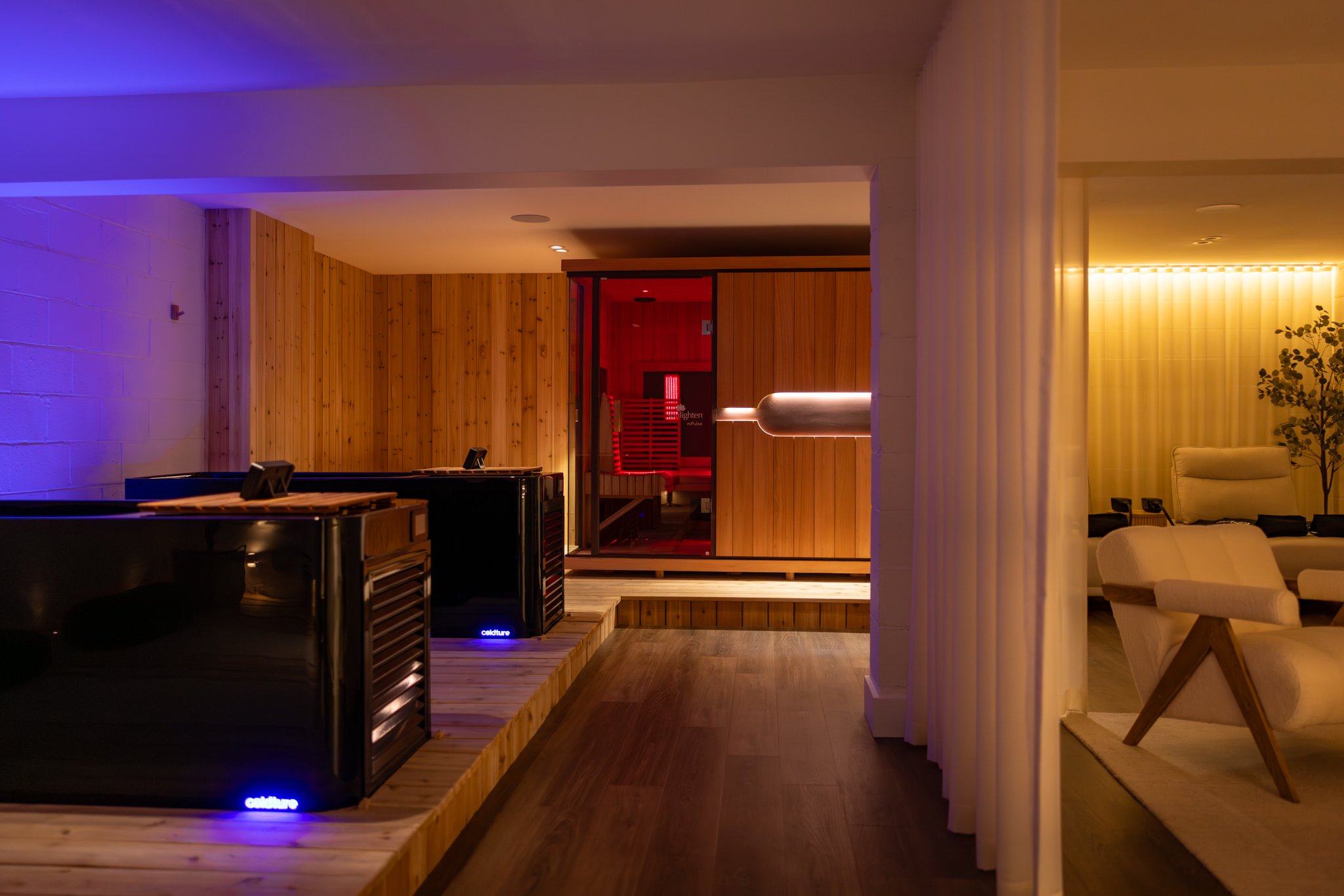 A cozy indoor lounge area with warm lighting, featuring a sauna with wooden walls and infrared light panels, next to a  fireside lounge area with cozy chairs.