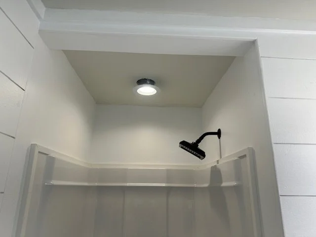 Empty shower with white walls and a black showerhead, ceiling light, and white door or wall panels.