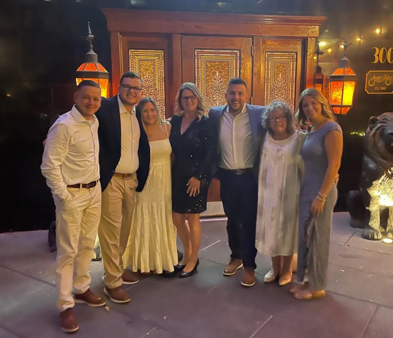 A group of eight adults standing together outdoors at night, posing for a photo in front of decorative wooden panels and illuminated lanterns, dressed in semi-formal attire.