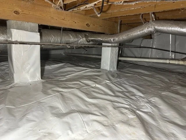 An unfinished basement with a vapor barrier on the floor, HVAC ductwork, and wooden beams overhead.