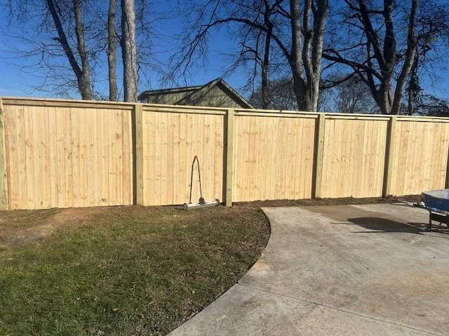 A wooden privacy fence in a backyard, with a scooter leaning against it, and a concrete patio area with a parked vehicle partially visible.