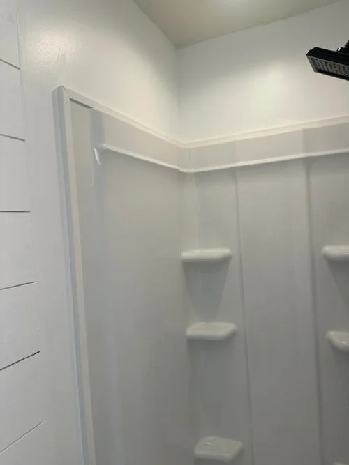 White corner shower stall with built-in shelves and a black showerhead in the top right corner.
