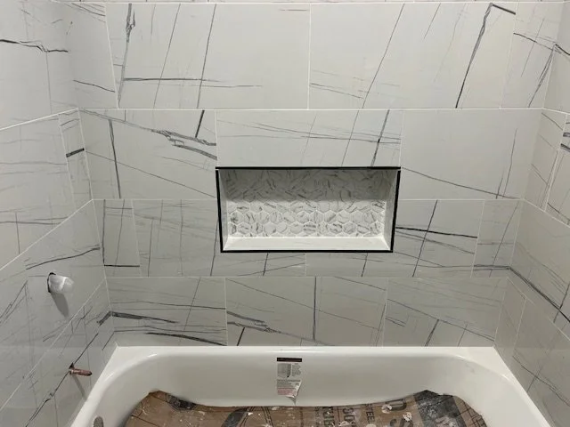 Bathroom wall with large marble tiles, a built-in niche with textured white stones, and part of a bathtub with a newspaper lining the bottom.