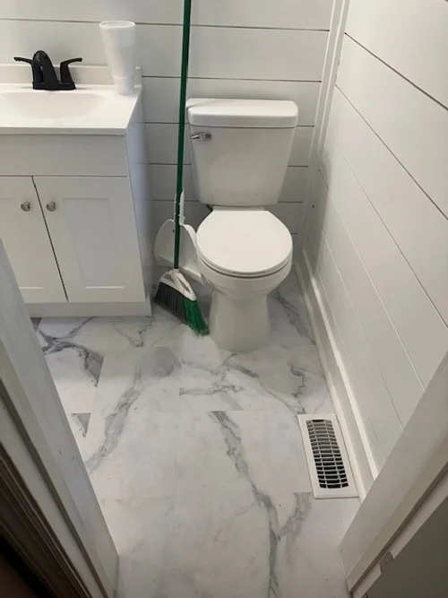 A small bathroom with a toilet, a white cabinet with a sink, a broom, and a flat vent on the marble floor.