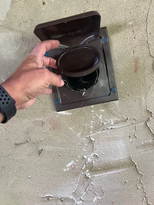 A hand lifting a black cover from a small, black outdoor electrical outlet mounted on a concrete wall.