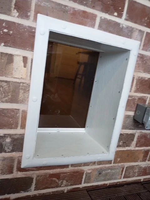 A white framed pet door with a transparent flap installed in a reddish-brown brick wall.