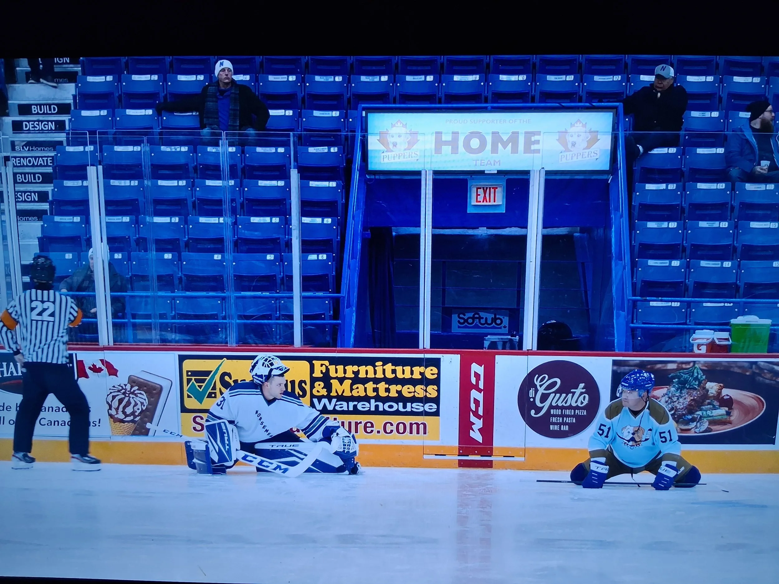 Hockey players and referee on ice rink with empty blue seats in the background, advertising boards along the side of the rink, and a few spectators seated in the stands.