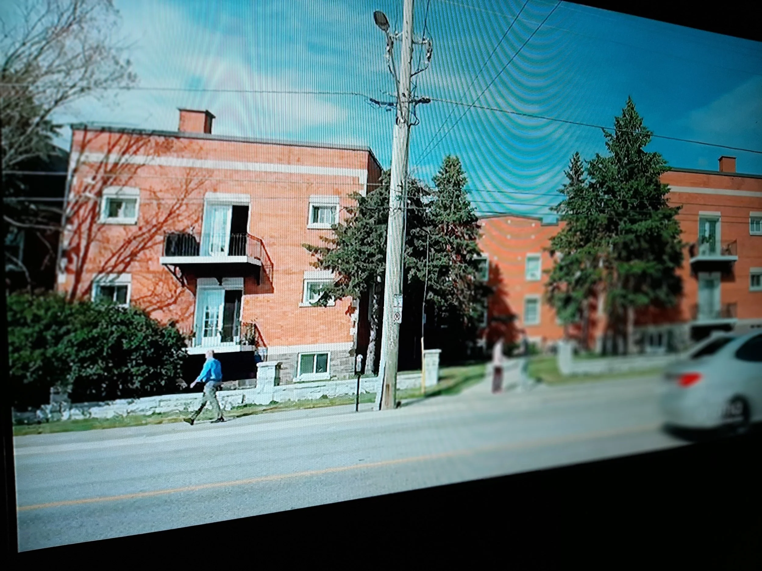 A street scene with two brick apartment buildings, trees, a utility pole, pedestrians, and a car.