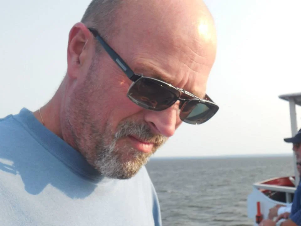 A bald man with a beard wearing sunglasses and a light blue shirt, standing outdoors near a body of water.